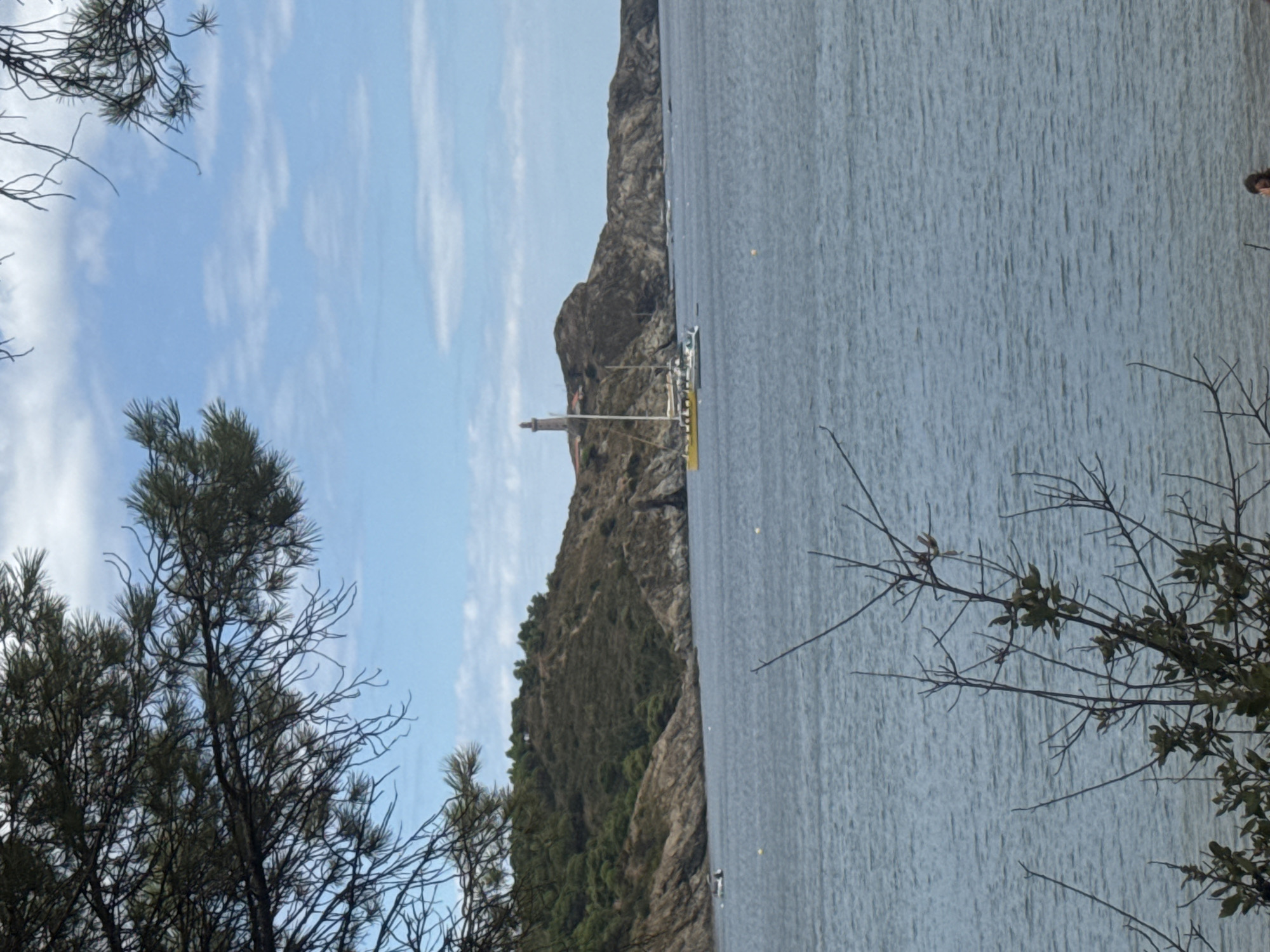 An outdoor, eye-level, medium shot captures a coastal scene in Port-Vendres, France, dominated by a body of water that occupies approximately two-thirds of the frame. The water is a muted blue, with a gentle ripple pattern across its surface. In the middle ground, a rocky, tree-covered hill rises from the water's edge. Perched atop this hill is a prominent lighthouse, its tall, slender structure clearly visible against the sky. In front of the hill, a yellow sailboat with white sails is anchored, its masts reaching towards the sky. A smaller, grey boat is positioned behind the yellow sailboat. To the left of the sailboat, a smaller white boat is visible. In the foreground, the branches of a dark, sparse tree with some leaves obscure parts of the scene, casting a shadow over the lower portion of the image. A small, blurry figure of a person is visible in the bottom right corner of the frame, near the water's edge. The sky above is a pale blue, dotted with scattered white clouds.