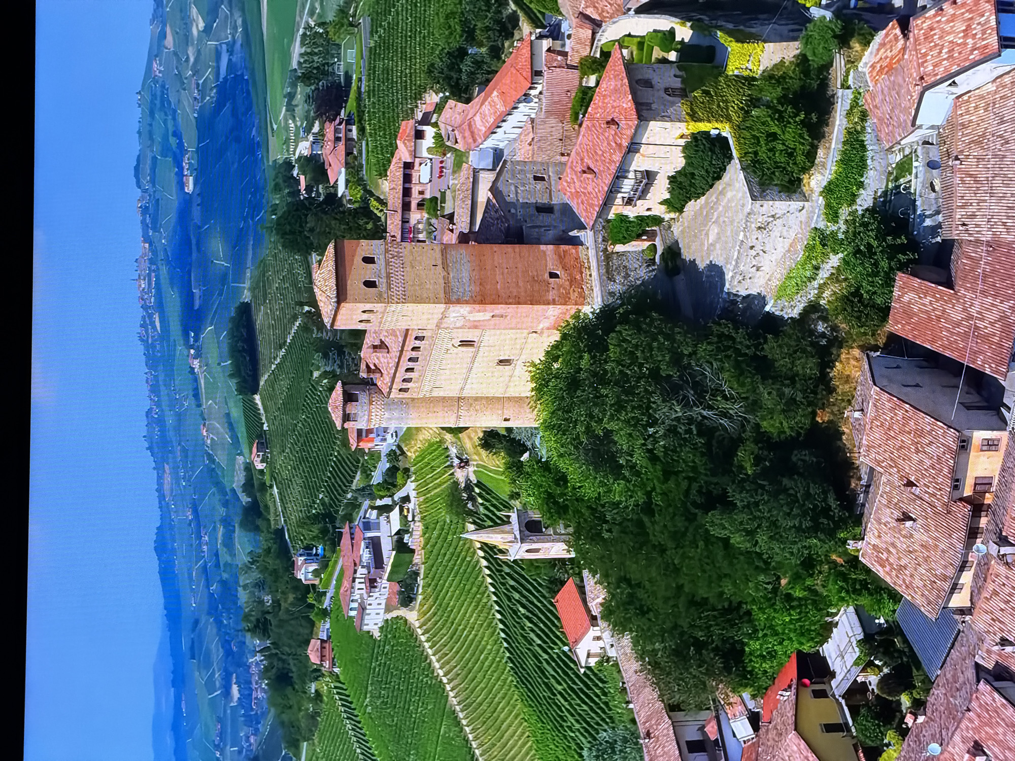 The image depicts a scenic aerial view of a European rural landscape, characterized by rolling hills covered in meticulously cultivated vineyards. A prominent medieval-style brick castle, featuring tall towers and arched windows, dominates the foreground. Adjacent to the castle are several other buildings with red tiled roofs, creating a village-like atmosphere. The surrounding terrain is lush with greenery, interspersed with patches of trees. The sky is a clear, bright blue. The scene evokes a sense of historical charm and agricultural abundance. The location is definitively not Chirchik, Uzbekistan, as the architecture and landscape are distinctly European, likely from a region known for winemaking, such as Italy or France.