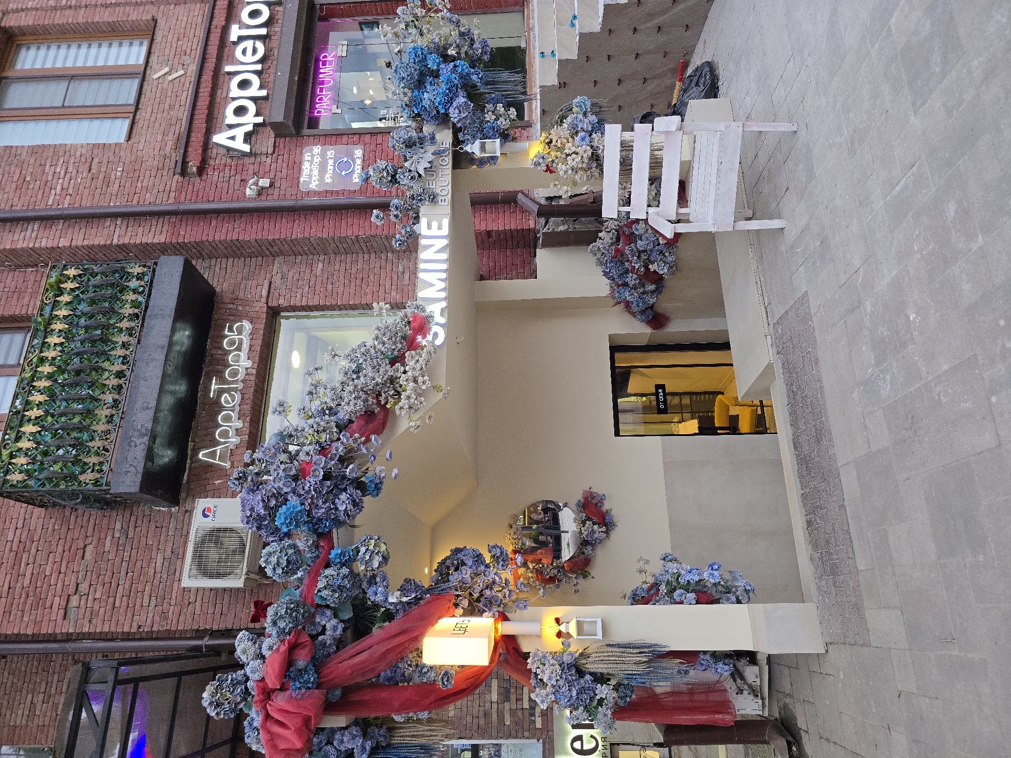 This image captures the storefront of a boutique called "SAMINE" located in an urban environment, likely in the city of Unknown, Unknown. The building's facade is a combination of brick and beige-colored walls, adorned with an elaborate arrangement of blue and white flowers, along with red fabric. To the left of the boutique is a store named "AppeTop95", with signage indicating it deals with Apple products (iPhone 15 and iPhone 16). "AppleTop" is also seen on another sign near the top of the image, along with the word "PARFUMER" on a neon sign. A white air conditioning unit is mounted on the brick wall, and an iron decorative piece is also visible. The entrance to "SAMINE" is partially obscured by the floral arrangement, leading into a warmly lit interior with chairs and tables visible through the doorway. A small, round mirror is placed on the upper wall. Outside, a white wooden structure adds to the decor, set against a paved sidewalk.