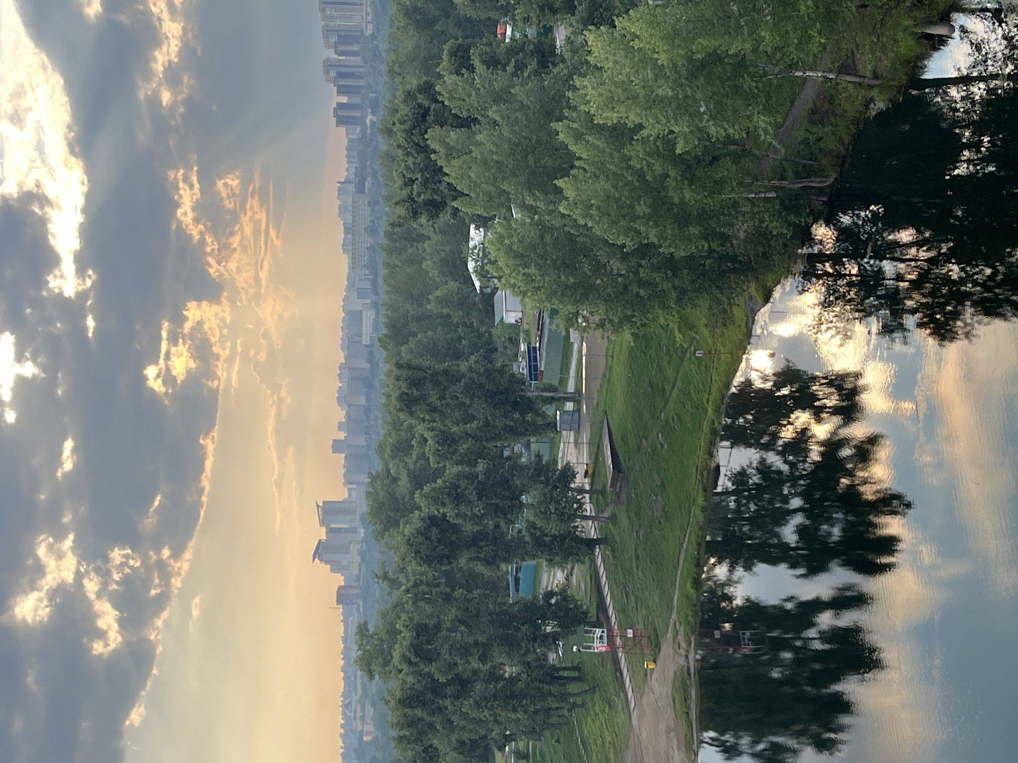 The image depicts a serene, wide-angle view of a park-like area with a body of water in the foreground. The sky is dramatic, with heavy, dark clouds illuminated by shafts of sunlight, suggesting either sunrise or sunset. The sun's rays pierce through the clouds, casting a golden hue on the upper atmosphere and reflecting onto the water.
In the foreground, a dark, calm body of water reflects the sky and the surrounding trees with remarkable clarity. The reflections create a mirror image of the natural elements, adding depth and symmetry to the composition.
Beyond the water, a lush green park unfolds. Numerous tall trees with dense foliage dominate the landscape, creating a natural canopy. Scattered among the trees are some man-made structures, including a small lifeguard-like tower with a red and white warning sign and a yellow trash bin, a few low-lying buildings with light-colored roofs, and some fences or barriers. A pathway winds through the grassy area.
In the mid-ground, a dense forest stretches across the scene. Further in the distance, a cityscape is visible, characterized by a cluster of modern, tall buildings. These buildings, with their geometric shapes and varying heights, stand in contrast to the natural environment, indicating the presence of an urban center.
The overall atmosphere is peaceful and picturesque, with a strong contrast between the raw beauty of nature and the constructed environment of the city. The lighting conditions suggest a specific time of day, contributing to the mood of the scene.
This scene is characteristic of the area along the Yenisei River in Krasnoyarsk, Russia, particularly park areas that offer views of the city skyline across the water. Specifically, this view is likely from the Karaulnaya Hill area, looking towards the northern part of the city, which features numerous residential and commercial high-rises visible from elevated viewpoints. The presence of a body of water and forested parkland is consistent with the topography and recreational spaces found along the riverbanks within or adjacent to the city.