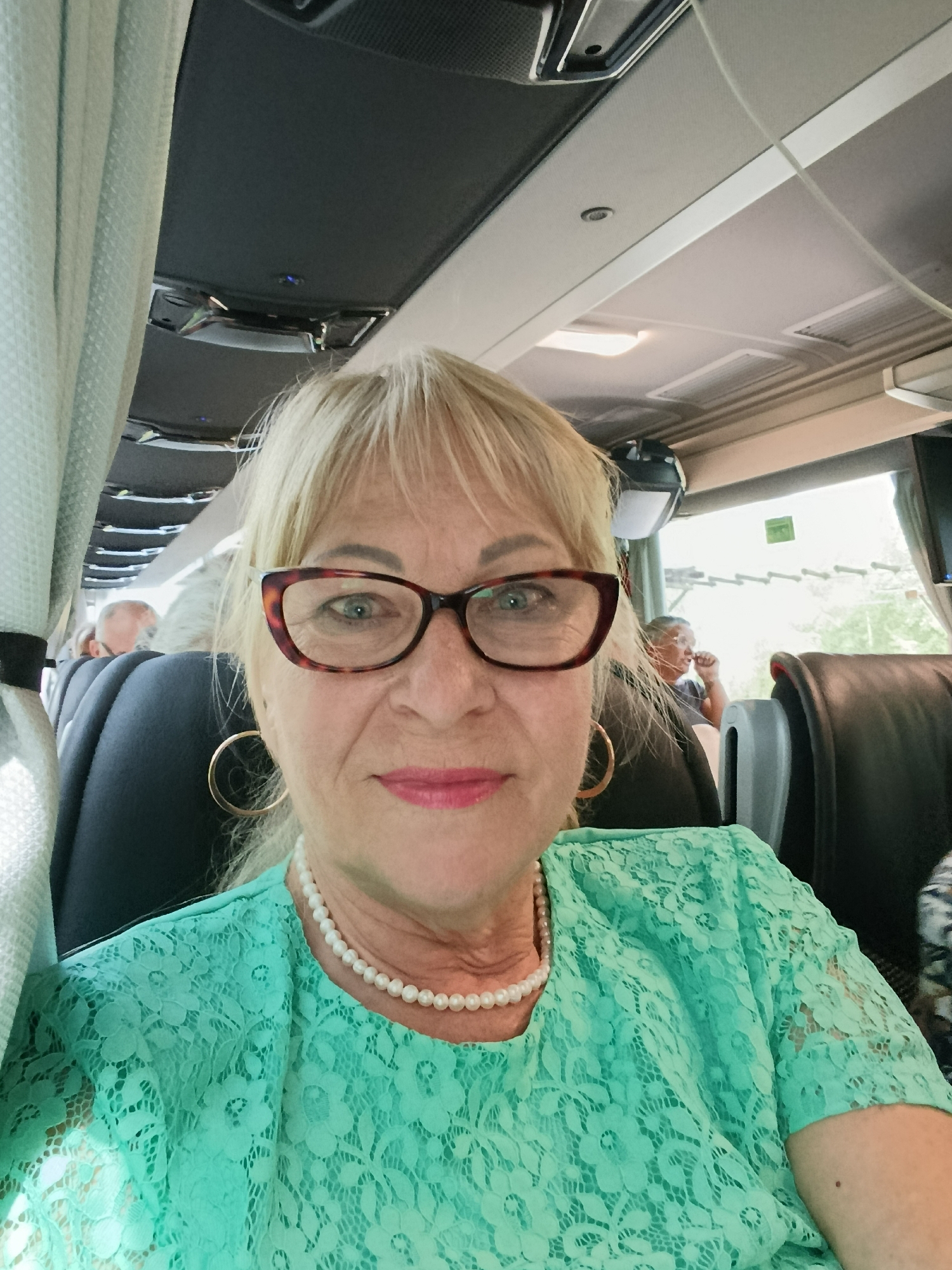 An elderly Caucasian woman with blonde hair and glasses is taking a selfie on a bus. She is wearing a teal lace top and a pearl necklace. The bus has black seats and a curtain on the left side. In the background, other passengers can be seen. The lighting suggests it is daytime. The image appears to be taken in Terras de Bouro, Portugal, as indicated by metadata.