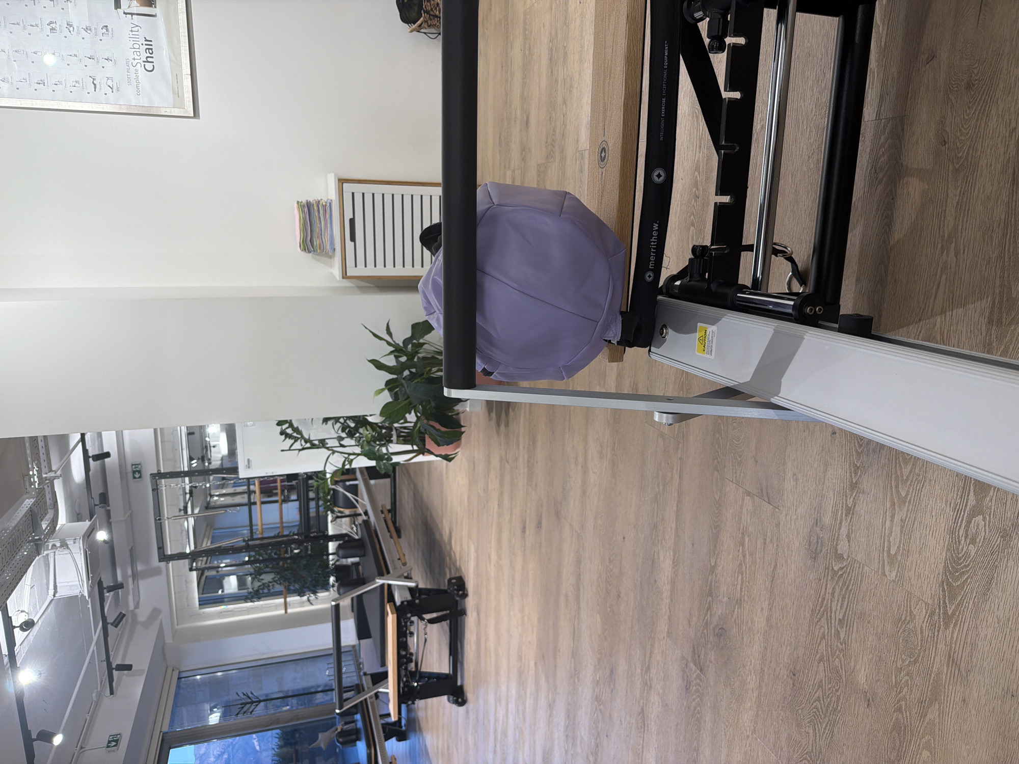 The image depicts the interior of a Pilates studio in Boreti, Montenegro. The scene is dominated by Pilates equipment, specifically reformers, with wooden floors and modern lighting fixtures. In the foreground, a portion of a reformer is visible, featuring a wooden carriage and metal components. A light purple duffel bag rests on the carriage. In the mid-ground, another reformer is positioned, and a stack of colorful towels is on a white cabinet. The background shows large windows with a view of the outdoors, a glimpse of plants, and a framed poster on the wall. The overall impression is one of a clean, well-equipped, and spacious fitness environment. There are no people visible in the image.