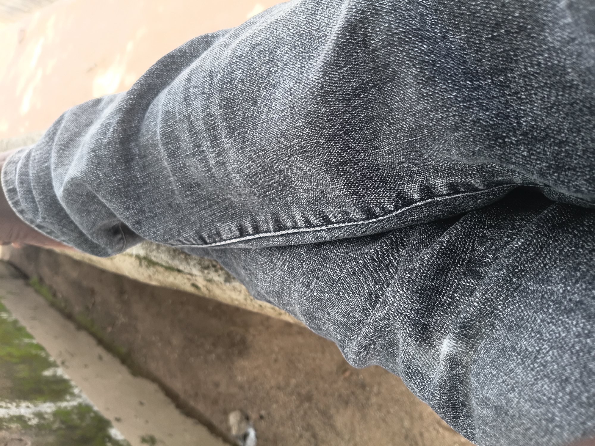 The image is a close-up shot of a person's legs, wearing dark gray denim jeans. The fabric of the jeans shows texture and some creasing, particularly around the knees and thighs, indicating movement or a seated position. The background is out of focus, but appears to be an outdoor setting with a concrete or stone ledge and a dirt or sand path. There is some greenery visible on the ledge in the upper left portion of the image. A small white object, possibly debris, is on the path. The lighting suggests it is daytime. Based on the context provided by the user, this scene is located in Jalingo, Nigeria.