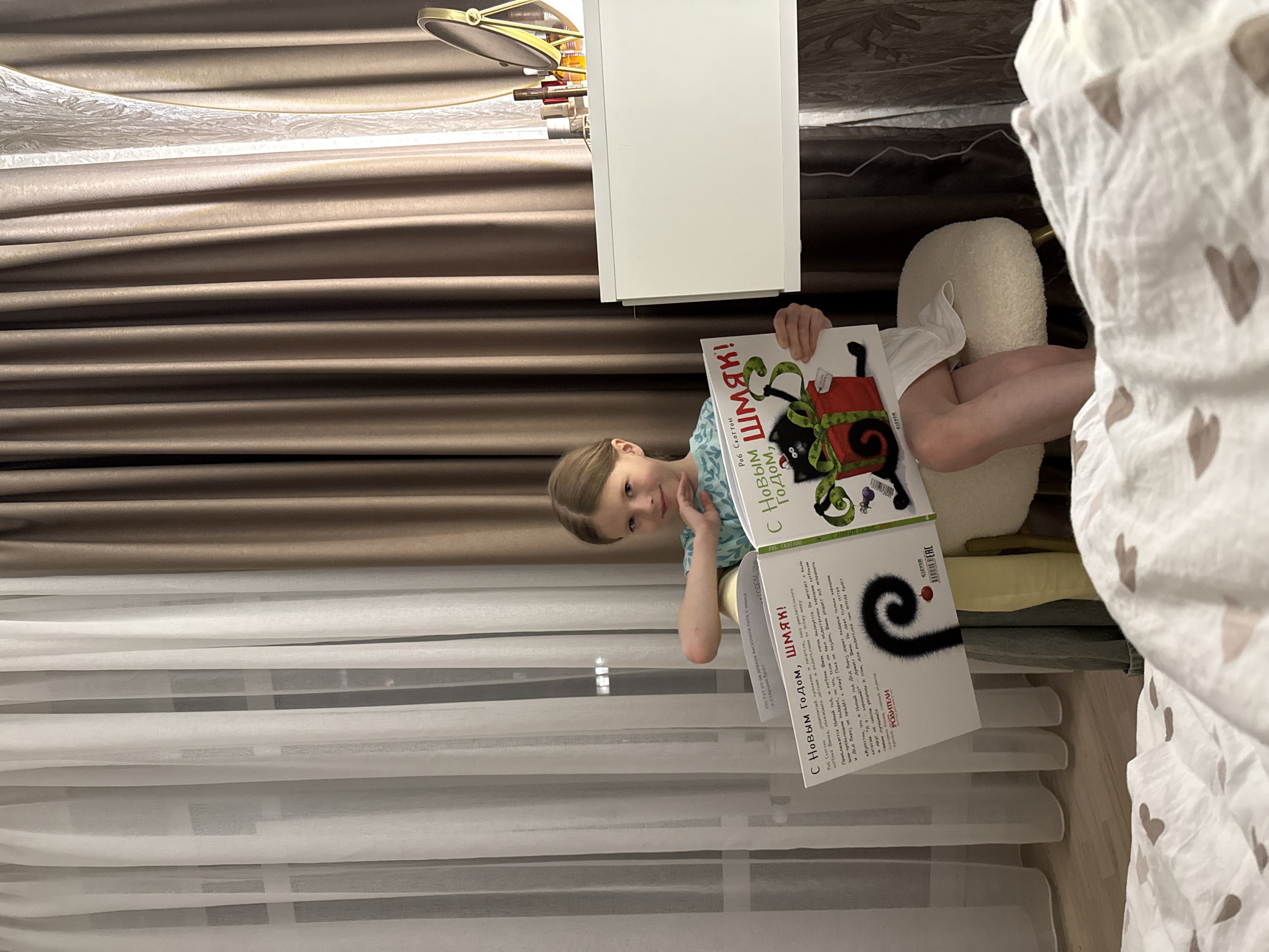 The image shows a young girl holding a book titled "Happy New Year, Shmyak!" in Russian. She is sitting on a white, tufted ottoman and is partially obscured by a white comforter with gray heart patterns. She is wearing a light blue patterned shirt. Behind her are brown curtains and a white dresser with shoes and other items on top. A white, sheer curtain is visible in the lower-left of the frame. Based on the book's text being in Russian, it's reasonable to assume the image was taken in a Russian-speaking region. Given that Izhevsk is in Russia, it is plausible, but not confirmed.