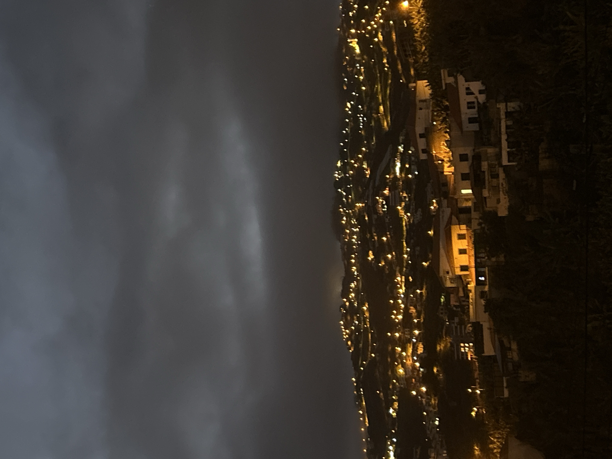 The image shows a nighttime view of Câmara de Lobos, Portugal. The sky is overcast and dark. The city is built on a hillside, with buildings illuminated by artificial lights. The lights appear as numerous small yellow points scattered across the slope. Some individual houses and structures are visible, with lit windows and outlines. To the right is a steep land, and to the left is a dark sky.