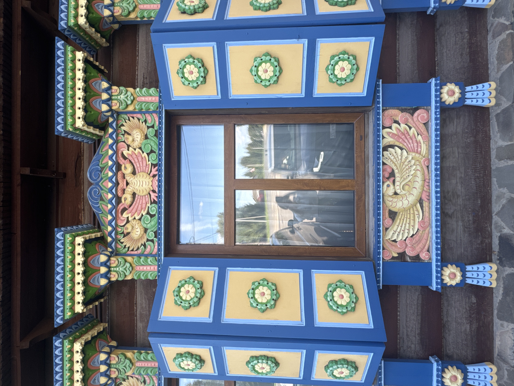 The image shows a close-up of the window of a traditional wooden house in Suzdal, Russia. The window is framed by intricately carved and painted wooden elements. The shutters are painted blue with yellow panels, each containing a green and yellow floral motif. Above the window, there is a decorative frieze with a depiction of a stylized bird, painted in pink, yellow, and green. The wooden walls of the house are visible below the window, with a stone foundation at the bottom. The window glass reflects the sky and trees.