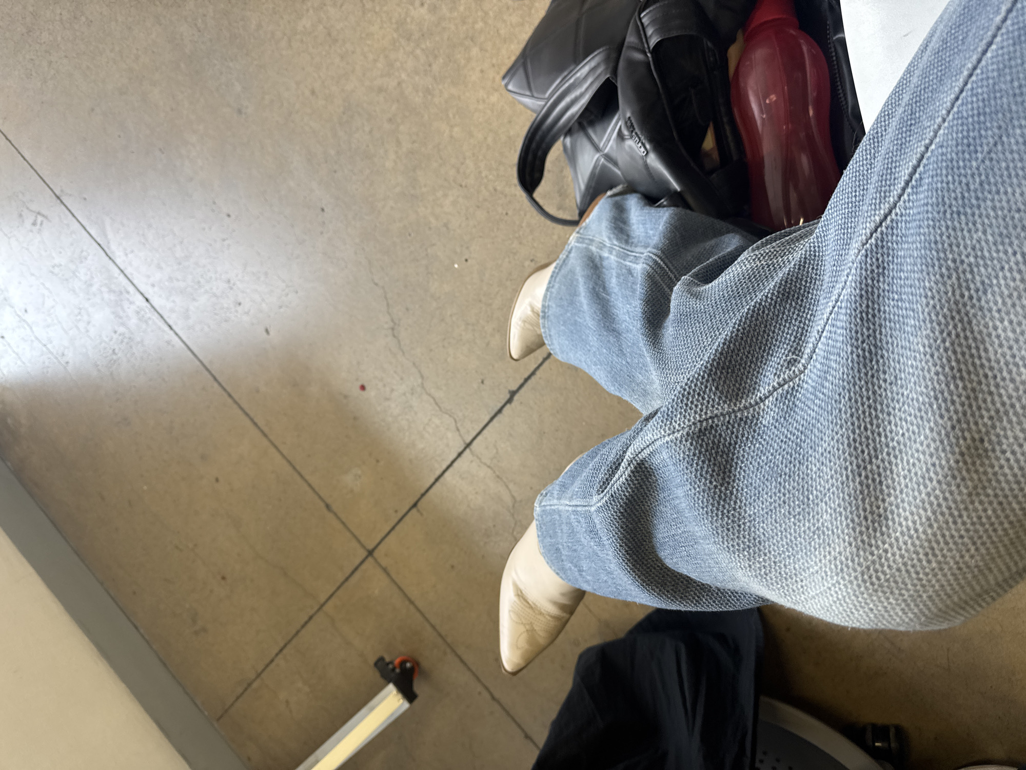 The image depicts a person seated, wearing light-wash denim jeans and beige pointed boots. Beside the person is a black bag with a red water bottle partially visible inside. The floor is a neutral tone, possibly concrete or tile, with linear patterns suggesting sections. A portion of a metallic object is visible at the bottom left of the frame. The setting appears to be indoors. The location cannot be definitively determined as Mexico City, Mexico, based solely on the visual information provided.