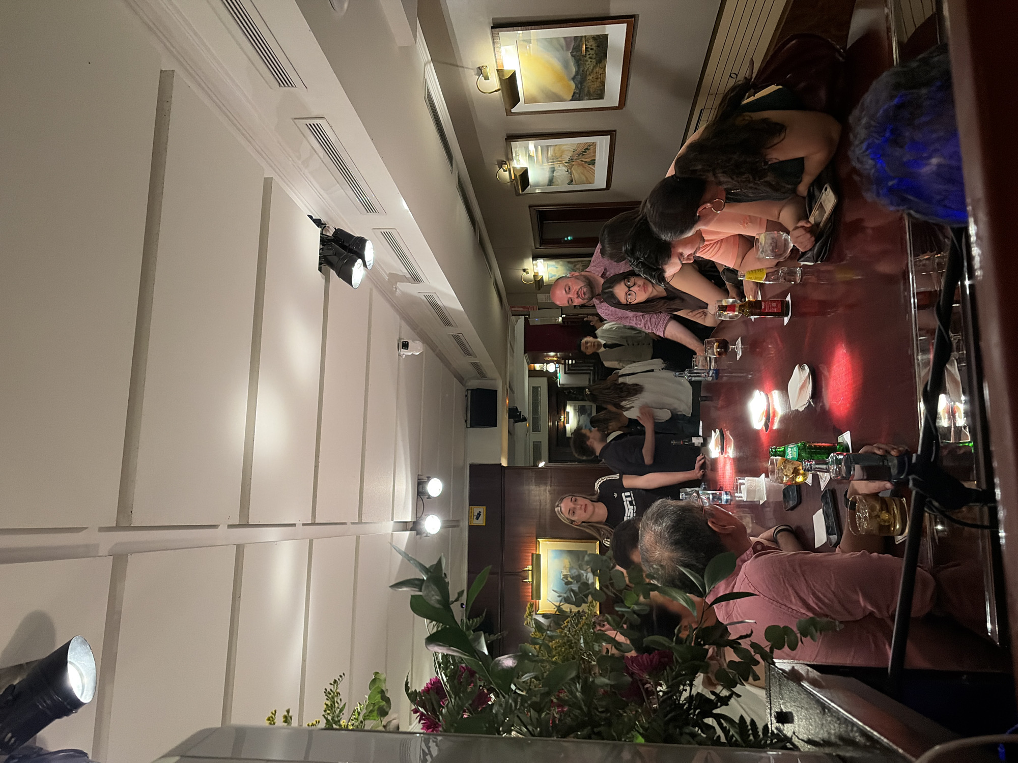 This image shows a group of people sitting at a long, dark wooden table in what appears to be a bar or restaurant setting. The location is in the city of Unknown, Unknown. Several people are conversing and interacting around the table, some are looking at their phones. Various drinks and glasses are visible on the table. The room is lit with overhead lighting, including track lighting along the ceiling, and sconces with framed artwork on the walls. In the foreground, a floral arrangement is visible. The wall features paneled construction.