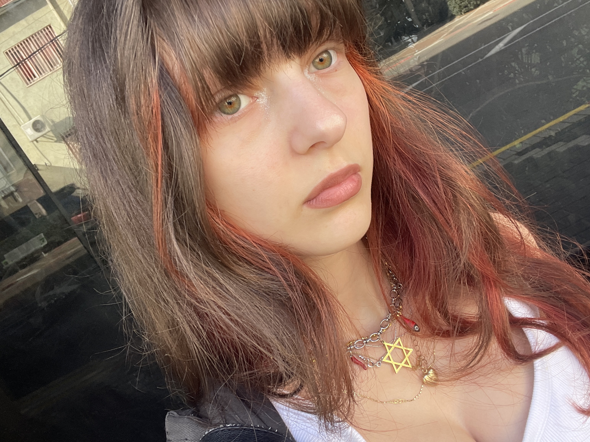 A young woman with brown hair, reddish-brown highlights, and bangs is looking directly at the camera. She has light skin, green eyes, and light pink lipstick. She is wearing a necklace with a Star of David pendant. She is wearing a white top and a jacket. The background is a street scene with buildings and vehicles, possibly in the city of Petah Tikva, Israel. The light suggests daytime.