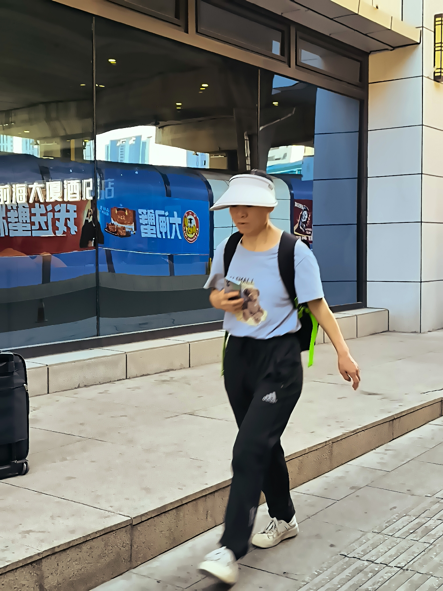 A person with a light blue t-shirt and black pants is walking on a sidewalk in front of a building with a large glass facade. The person is wearing a white visor and a backpack with green straps. They are holding a smartphone in their right hand. To the left of the person, a black suitcase is visible. The glass facade reflects a blue banner with Chinese characters and images. The sidewalk is made of light-colored concrete slabs, and there is a raised curb separating the sidewalk from the street. The building has a modern architectural style with white tiled walls and horizontal windows.