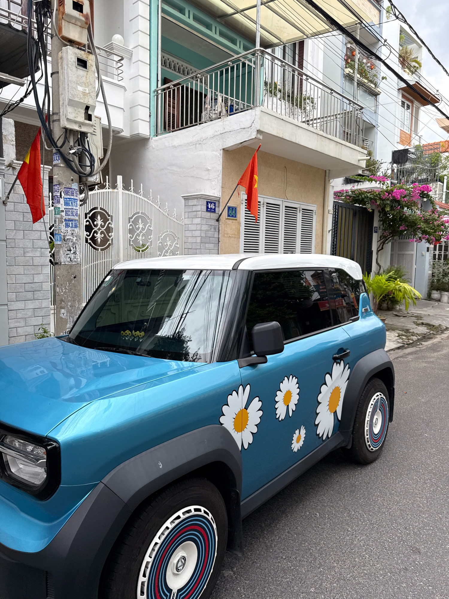 A blue and white car with daisy decals is parked on a residential street in Nha Trang, Vietnam. The car is a modern electric vehicle with a white roof and black trim. The daisy decals are large and white with yellow centers, and they are arranged on the side of the car. The car is parked on the right side of the street, and there is a sidewalk and a building behind it. The building is a two-story house with a balcony on the second floor. The house is painted white and has a light blue trim. There is a red and yellow flag hanging from the balcony. The street is paved with asphalt, and there are some trees and plants on the side of the street. The sky is overcast, and the light is diffused. The scene appears to be peaceful and quiet.
