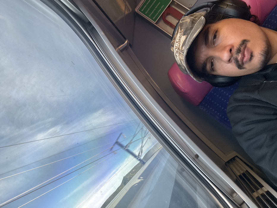 A man wearing a camouflage hat and headphones reclines against a seat with pink and blue upholstery. He has a mustache and a light beard. The scene is set inside a train carriage, as evidenced by the window reflecting an exterior view of power lines and land. A green sign with white lettering and a red metal object are visible above the seat. The overall composition is taken at an angle, emphasizing the window and the man's relaxed posture.