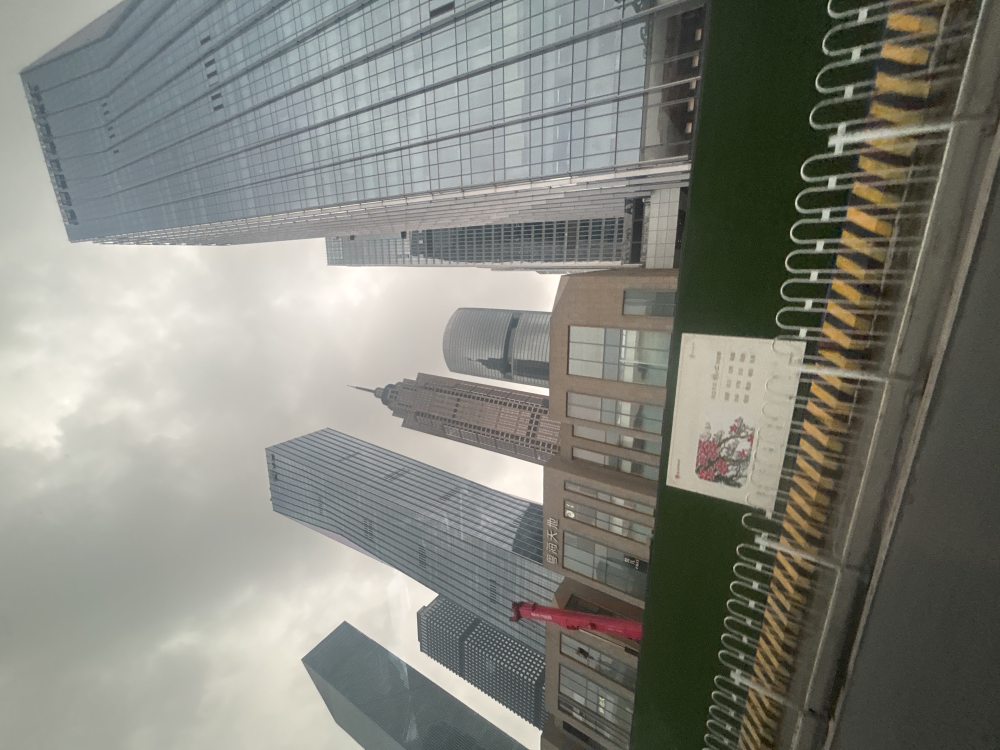 This is a ground-level, wide-angle shot of a city skyline in Tianhe District, China, under an overcast sky. The focus is on a cluster of modern skyscrapers of varying heights and architectural styles. On the right, a very tall, glassy skyscraper dominates the frame, reflecting the sky. To its left, another tall, slender skyscraper with a patterned facade is visible. Further left, a building with a distinctive stepped, tiered design stands out, followed by a dark, angular skyscraper. In the foreground, a low-rise building with a beige facade and large windows is partially obscured by a green construction barrier. A bright red, abstract sculpture is visible against the beige building. A white metal fence with yellow and black striped barriers runs along the bottom of the frame, separating the road from the construction site. A white sign with Chinese text and an image of red flowers is mounted on the fence. The street in the foreground is paved and appears wet, suggesting recent rain. There are no people visible in the image.