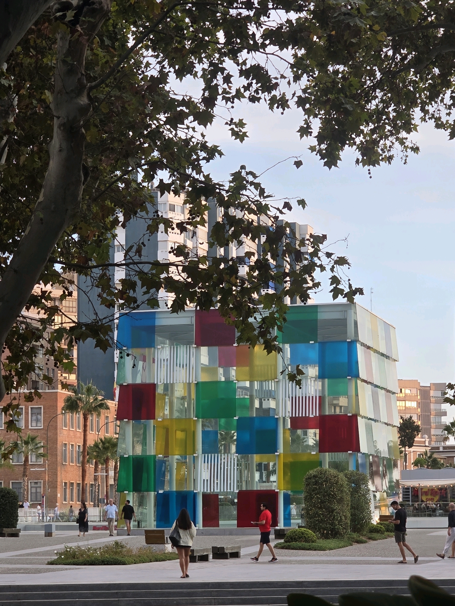 The image depicts the Centre Pompidou Málaga, a distinctive cultural center situated in the port of Málaga, Spain. The building's facade is characterized by its vibrant, multi-colored glass panels in shades of red, blue, green, and yellow, creating a striking, modern aesthetic. The structure is a geometric assemblage of cubes, with some panels appearing opaque and others transparent, revealing the interior.
In the foreground, a large tree with dark green leaves partially obscures the view of the building and the sky. The ground is paved with light-colored stones, and several people are visible walking across the plaza. To the left, a woman in a black dress and a man in a light blue shirt and khaki shorts are walking away from the camera. To the right, a woman in a striped top and skirt is walking towards the building, and a man in a red shirt and dark shorts is walking towards the left. Further in the background, other individuals are seen strolling, and palm trees add a touch of tropical vegetation to the urban landscape. Behind the multi-colored building, there are several other multi-story buildings, some with a modern architectural style and others appearing older with brick facades. The sky is a pale blue, indicating either early morning or late afternoon light. The overall scene suggests a public space within a city, likely a tourist or recreational area, given the presence of the cultural center and the casual attire of the people.