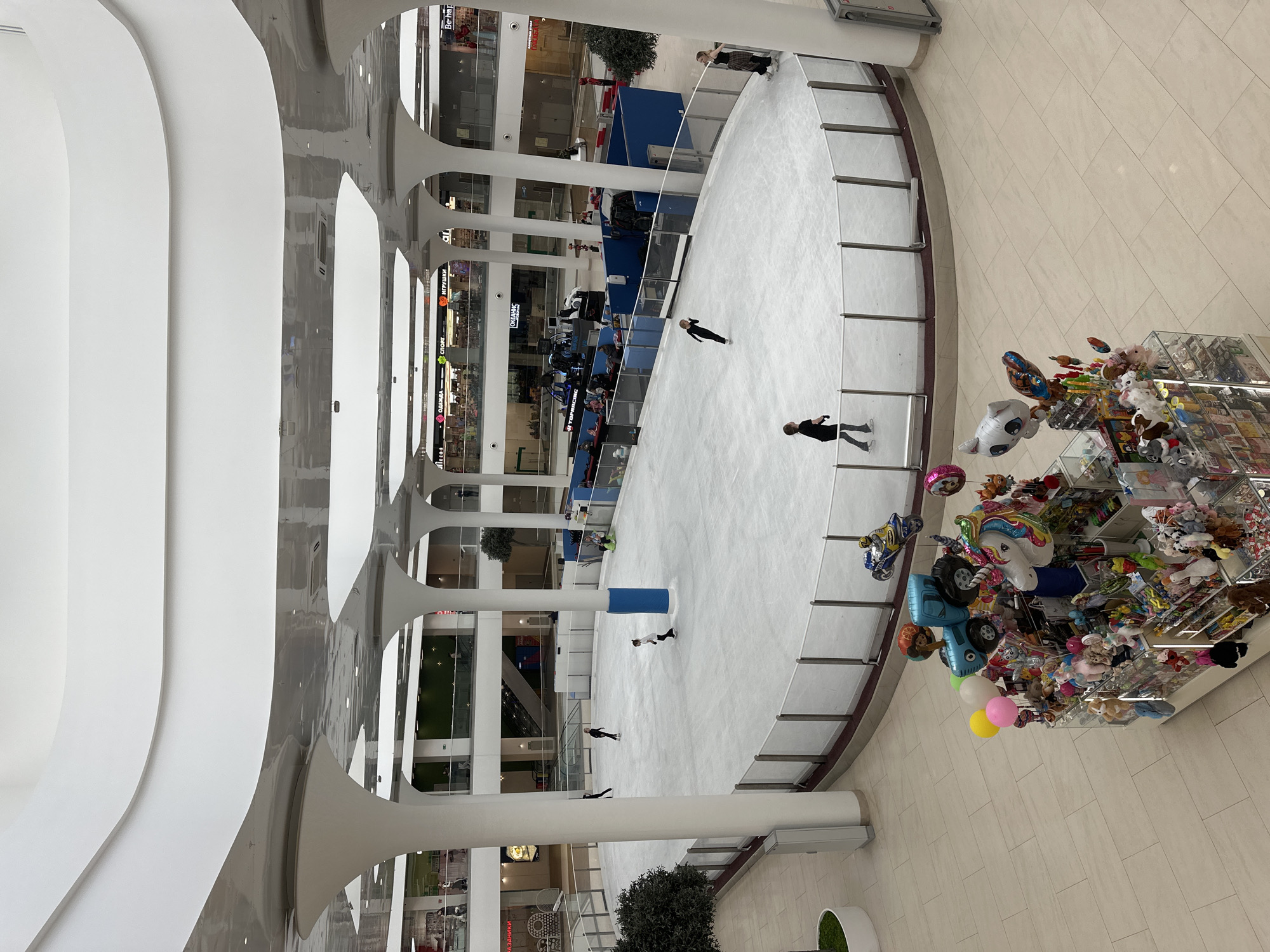 The image is a high-angle view inside the Seventh Heaven Shopping Center in Nizhny Novgorod, Russia. An indoor ice rink occupies the central space, where several people are ice skating. The rink is surrounded by a glass barrier. Adjacent to the rink, a merchandise kiosk displays balloons and toys. The shopping center's architecture features white columns and curved ceilings. The environment appears clean and well-lit.