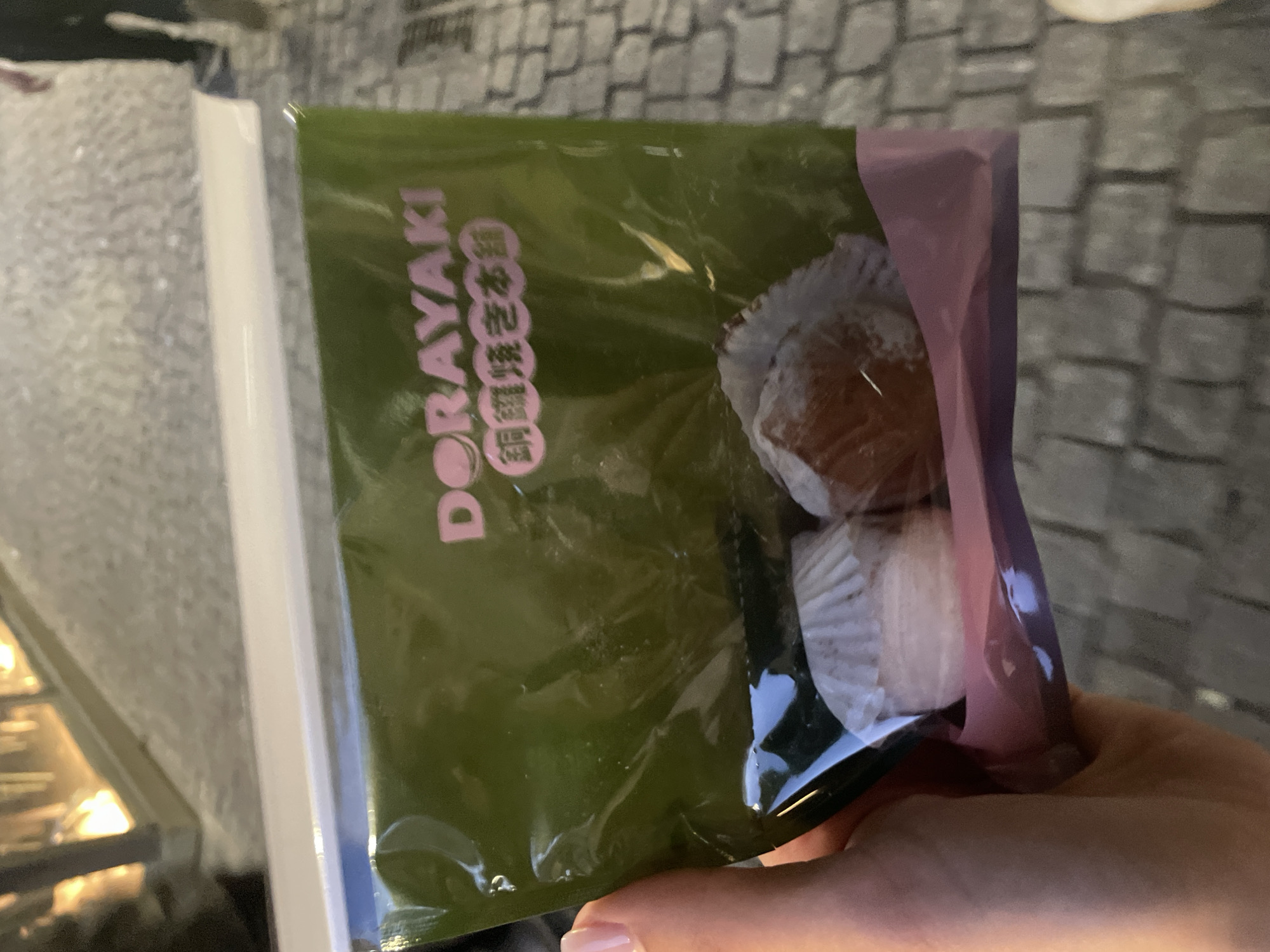 A person holds a packaged "Dorayaki" snack. The packaging is green with pink text and a pink border. The dorayaki is visible inside the package and appears to be in a white paper liner. The image appears to be taken indoors, with a blurred background suggesting a store setting. The location is possibly Milan, Italy, based on contextual clues.