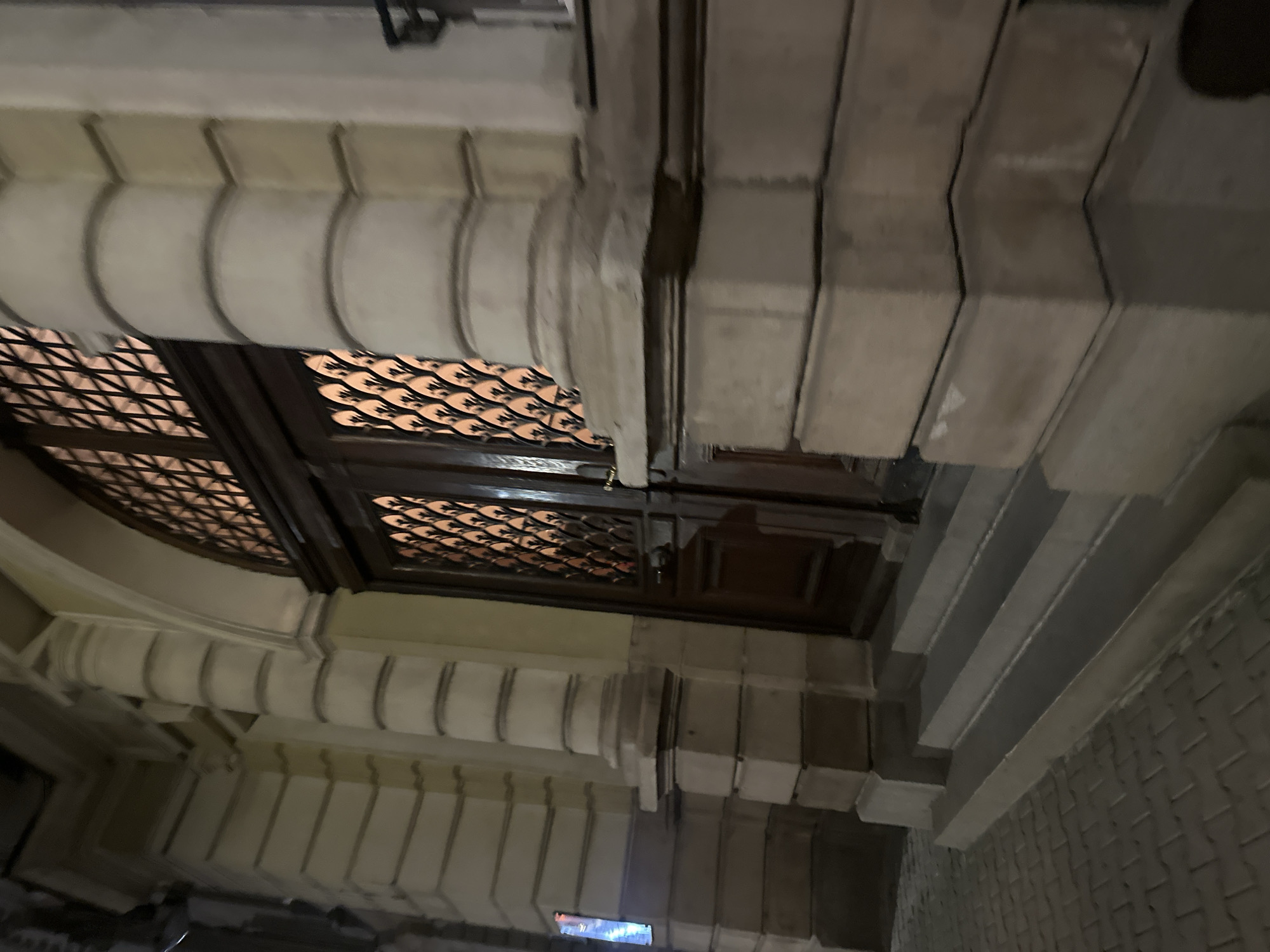 The image captures an exterior view of a building entrance at night. A grand wooden double door is centrally located, framed by a large arched window with a decorative metal grate. The door itself is paneled and features a round doorknob on the right side. The entrance is approached by a set of concrete steps that lead up from a brick-paved sidewalk. The building's facade is made of stone, with prominent columns and pilasters adorned with architectural details. The lighting suggests it is nighttime, with warm illumination emanating from within the building and casting shadows on the exterior. The scene is static and does not depict any individuals or active events. The location is described as "Unknown, Unknown."