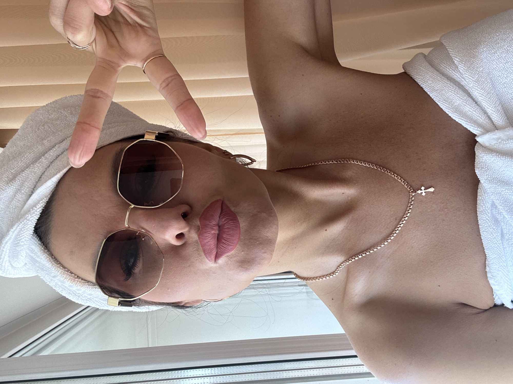 A woman with light brown skin is posing indoors, wearing sunglasses and a towel on her head, and a white towel around her body. She is also wearing a gold necklace with a cross. She is holding up a peace sign with her hand. The location is unknown.