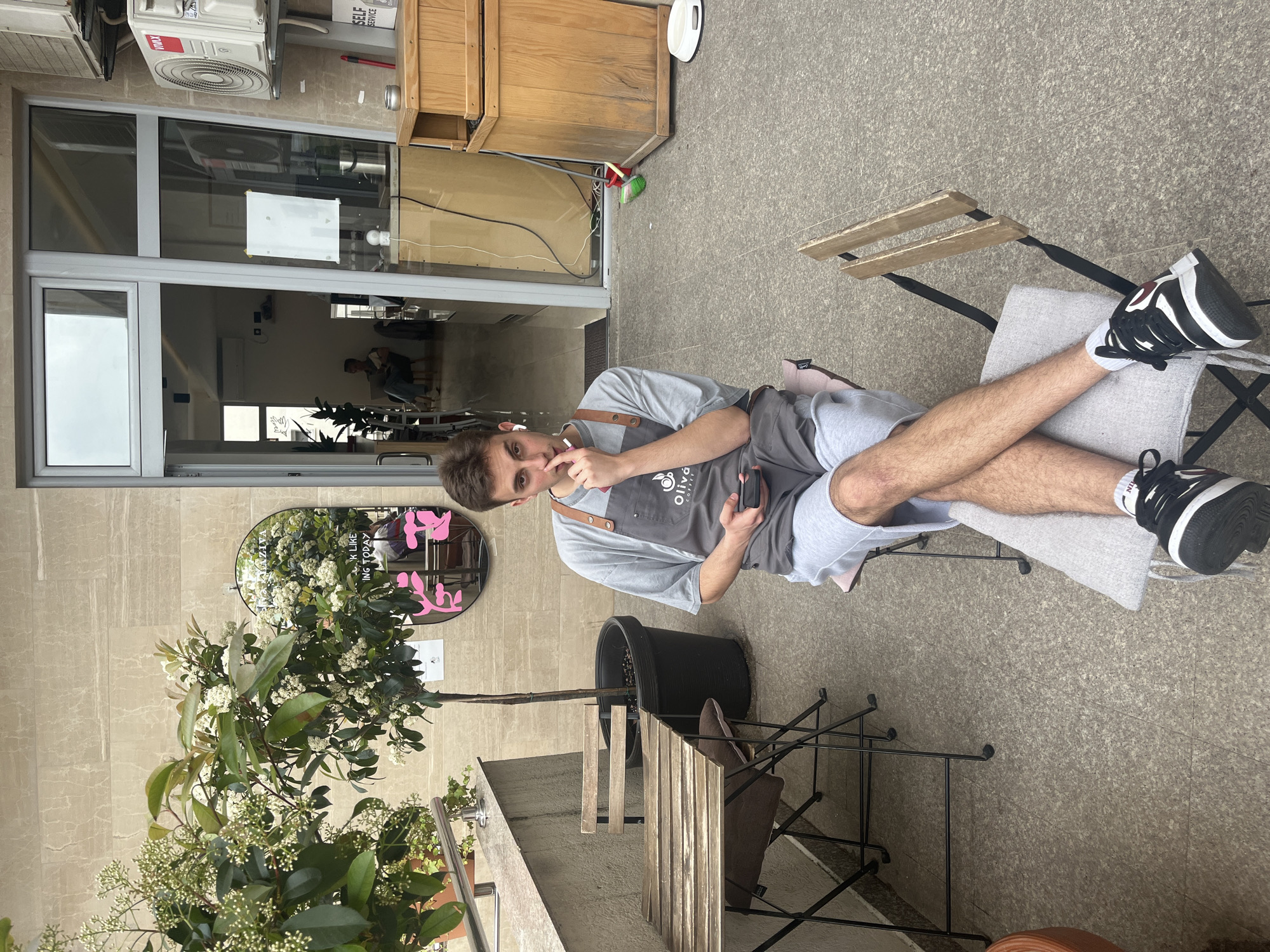 An outdoor, slightly elevated shot captures a young man relaxing on a cafe patio in Budva, Montenegro. The man, positioned centrally and looking directly at the camera, sits on a grey-cushioned chair with his legs crossed. He wears a grey t-shirt, grey shorts, black and white sneakers, and a grey apron with "Olivá Coffee" partially visible as a logo. He has earbuds in, holds a smartphone in his left hand, and brings a small pink object (possibly a vape pen) towards his nose/mouth with his right hand. His expression is neutral to slightly amused.
The setting appears to be a modern cafe patio with grey speckled pavement. To the man's right, there's a black metal table with wooden slats. A large potted tree with white flowers and green leaves stands between two tables. An oval mirror on the tiled wall behind the tree reflects parts of the scene and displays a pink graphic along with partially visible text like "LOOK LIKE ING TODAY."
The background shows the cafe's entrance, featuring a glass door and window reflecting the interior, where another person can be faintly seen. Above the entrance, an air conditioning unit with "VMAX" written on it is mounted on the wall. To the right of the door is a wooden cabinet-like structure, near which a white round object and a red brush handle are visible on the ground. The lighting suggests it is daytime, possibly overcast, as there are no harsh shadows. The overall scene conveys a casual, relaxed atmosphere at an outdoor cafe.