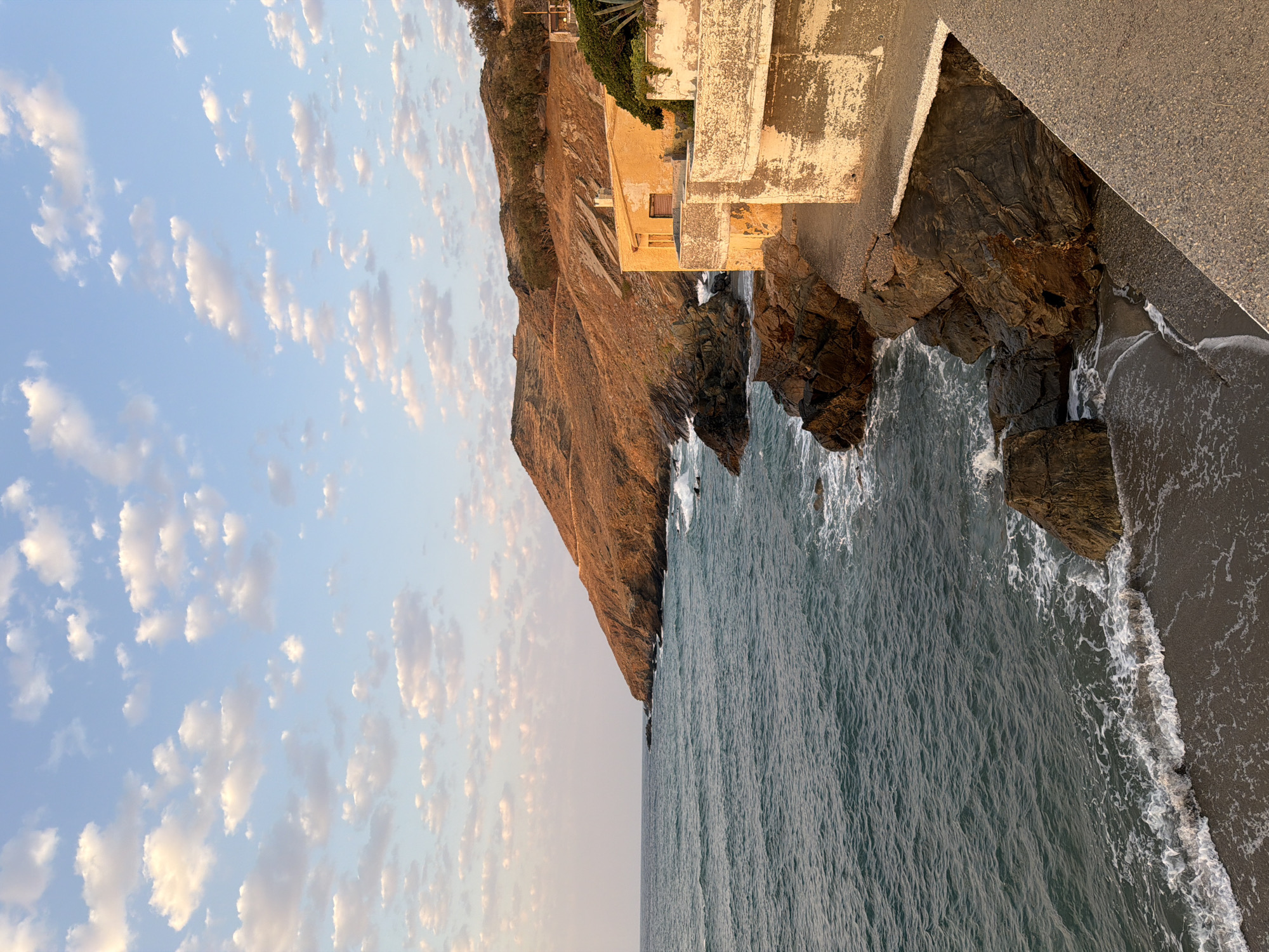 A vertical, eye-level, medium shot captures a coastal scene in Panormos, Greece, bathed in the warm, golden light of sunset. The turquoise sea gently laps against a rocky shoreline and a small strip of dark sand. White foam swirls in the shallow water as waves break against the rugged rocks.
Dominating the right side of the frame is a weathered, two-story building with faded yellow walls. Its architectural style suggests an older construction, with visible signs of aging and peeling paint. A concrete walkway, textured with small pebbles, slopes down from the foreground, leading towards the building and the sea. Behind the building, the terrain rises into a rugged, arid hillside covered in dry, scrubby vegetation. The distant hills, bathed in the setting sun's glow, appear a warm, reddish-brown. The sky above is a pale blue, dotted with scattered, fluffy white clouds that are beginning to take on hints of pink and orange from the sunset. The overall atmosphere is one of serene, late-afternoon tranquility.