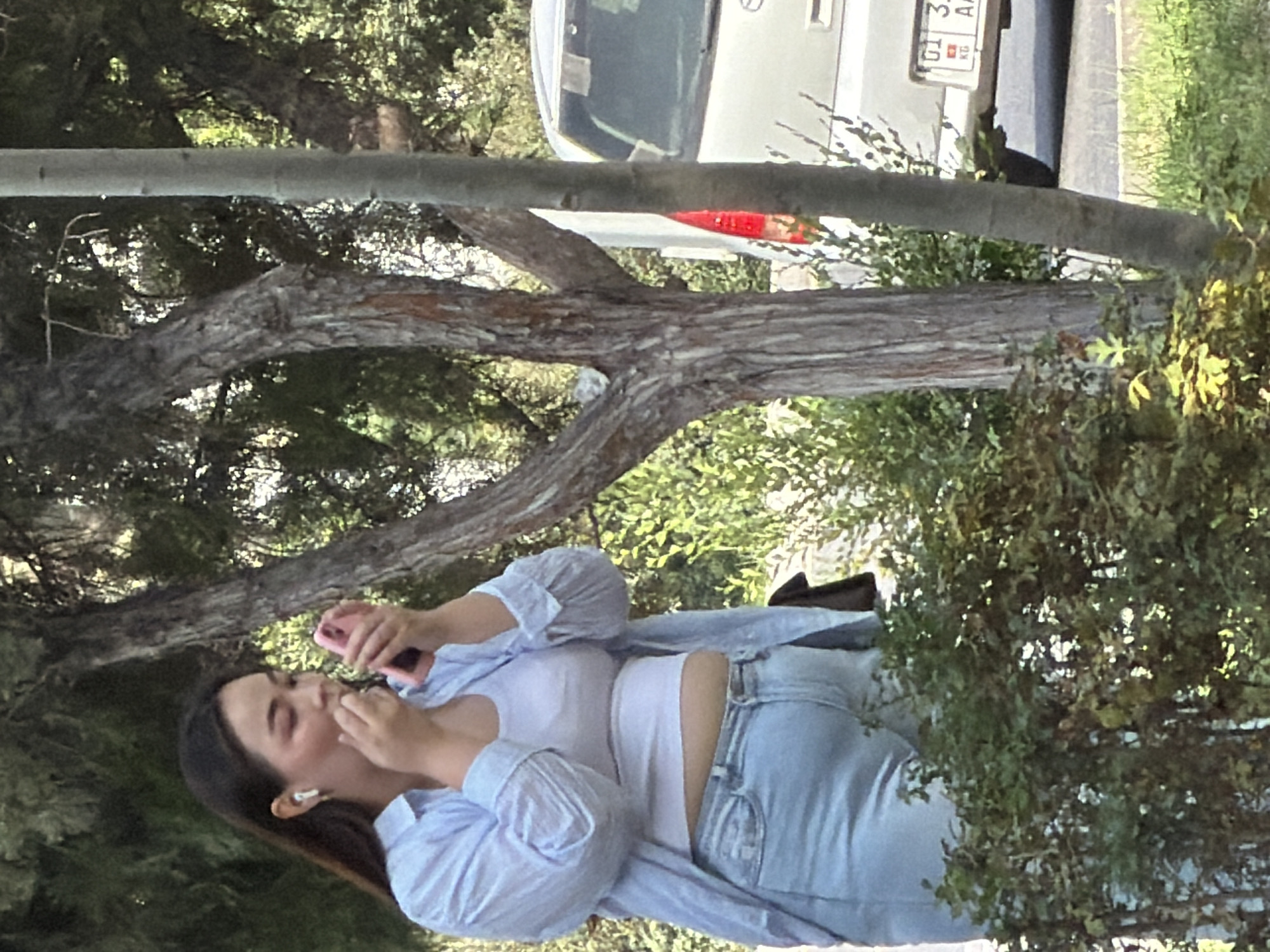 A woman with long dark hair and earbuds in her ears is applying makeup with a pink lipstick or gloss, holding a pink compact in her left hand. She is wearing a light blue open button-down shirt over a white tank top and light blue jeans. The woman is standing outdoors in a park-like setting with trees and bushes. In the background, a white van is parked on the side of a road. The license plate of the van is partially visible and appears to be from Kyrgyzstan.