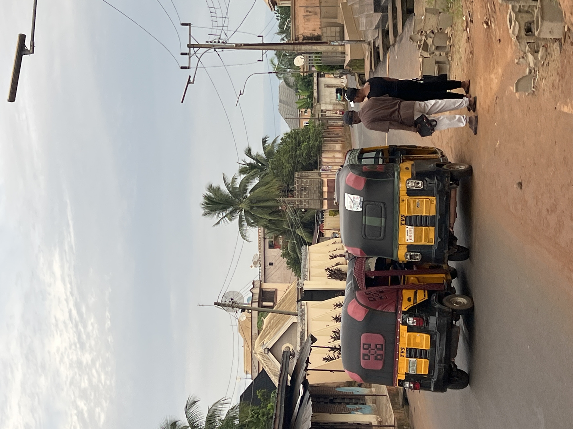 The image captures a street scene in Nkpor, Nigeria. Two yellow and black auto-rickshaws, also known as Keke Napeps, are parked side-by-side on a dirt and asphalt road. The auto-rickshaws are decorated with red and black patterns on their canopies. Three individuals are visible; two men stand behind the auto-rickshaws, facing away from the camera, with one man wearing a brown shirt and white pants, holding a bag, and the other wearing a black shirt and black pants. A third person, a man, stands further down the road to the left, partially obscured by the auto-rickshaws. Buildings, including houses and shops, line the street, with palm trees and electrical wires overhead. The sky is overcast, with scattered clouds. The scene appears to be in a residential or commercial area.