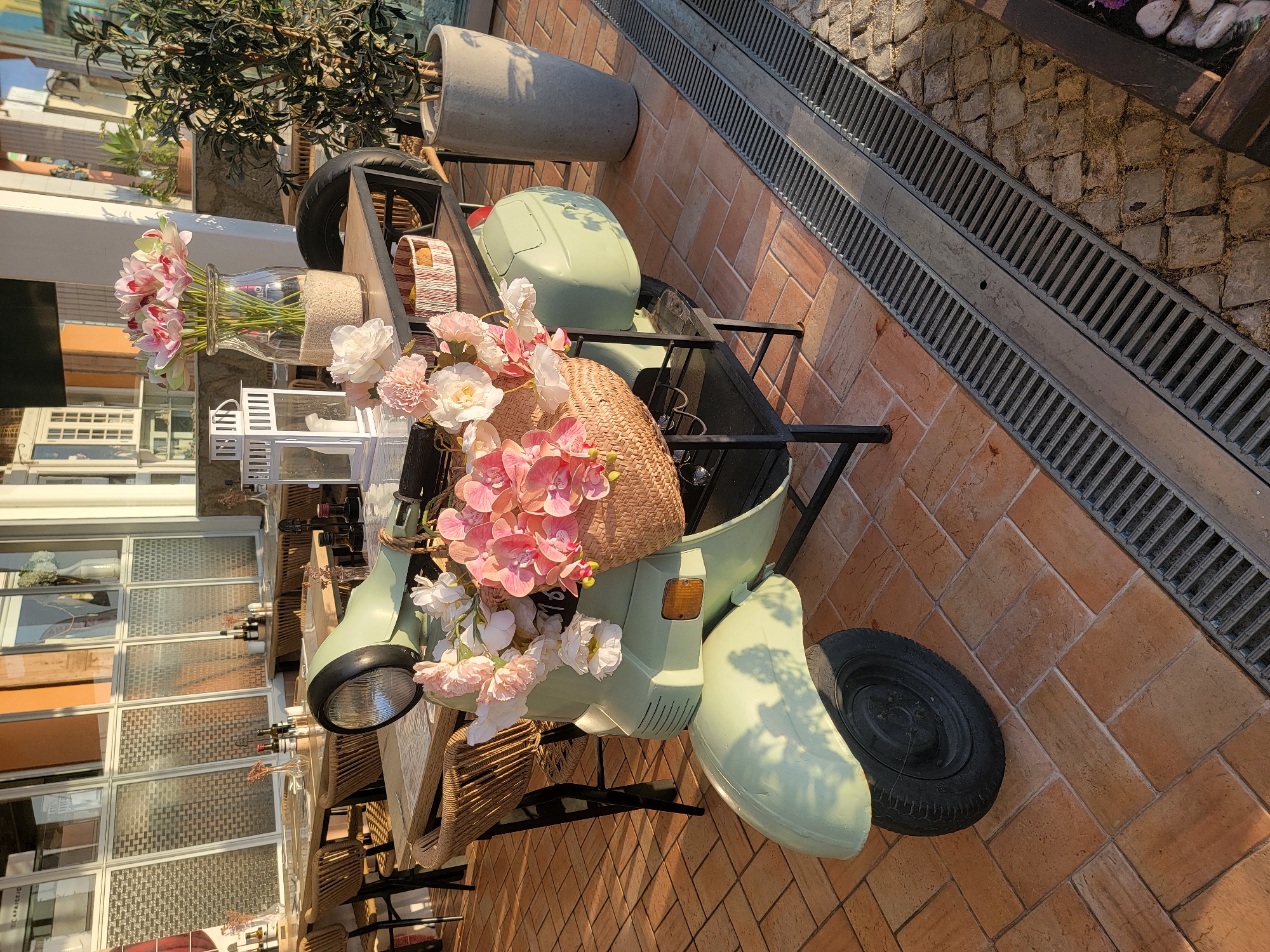 An outdoor seating area at a restaurant, possibly in Quarteira, Portugal, features a vintage-style light green scooter adorned with a straw basket full of pink and white artificial flowers. The scooter is parked on a tiled patio beside a table set with tableware and wine bottles, and a tall, grey concrete planter holding an olive tree. In the background, a building with large windows and outdoor dining tables creates a setting of a sunny afternoon.