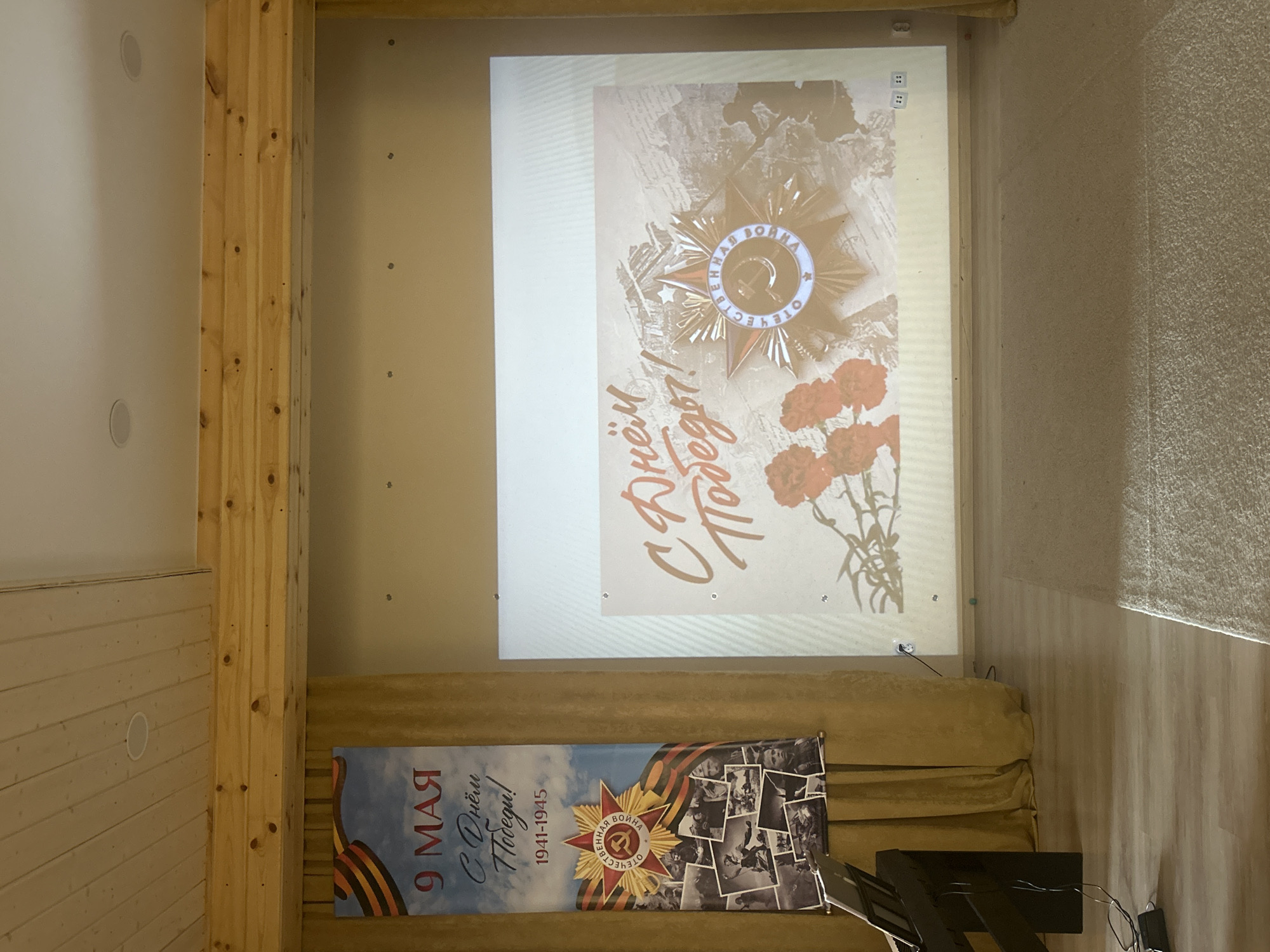 The image shows a room in Izhevsk, Russia, decorated with Soviet/Russian military-themed banners and projections, presumably for Victory Day (May 9th). A projector displays an image of the "Order of the Patriotic War" and carnations with the Russian text "С Днём Победы!" ("Happy Victory Day!"). A banner hanging below features a similar theme with the date range "1941-1945" and a montage of historical photographs. There is a wooden beam on the left and light walls on the right. A piece of audio equipment (likely a speaker) is visible in the bottom right. A gold colored couch is below the projections.