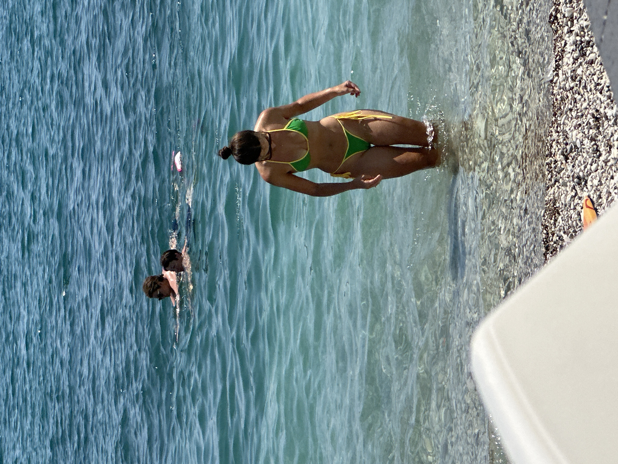 In the city of Budva, Montenegro, on a sunny day, a woman in a green and yellow bikini walks in the shallow, clear turquoise waters of the Adriatic Sea. Her hair is pulled up in a bun. In the background, two men are swimming further out in the sea, one wearing sunglasses. The seabed is covered in small, light-colored pebbles, visible through the water. The foreground shows a white object, possibly part of a lounge chair, and a patch of the pebble beach.