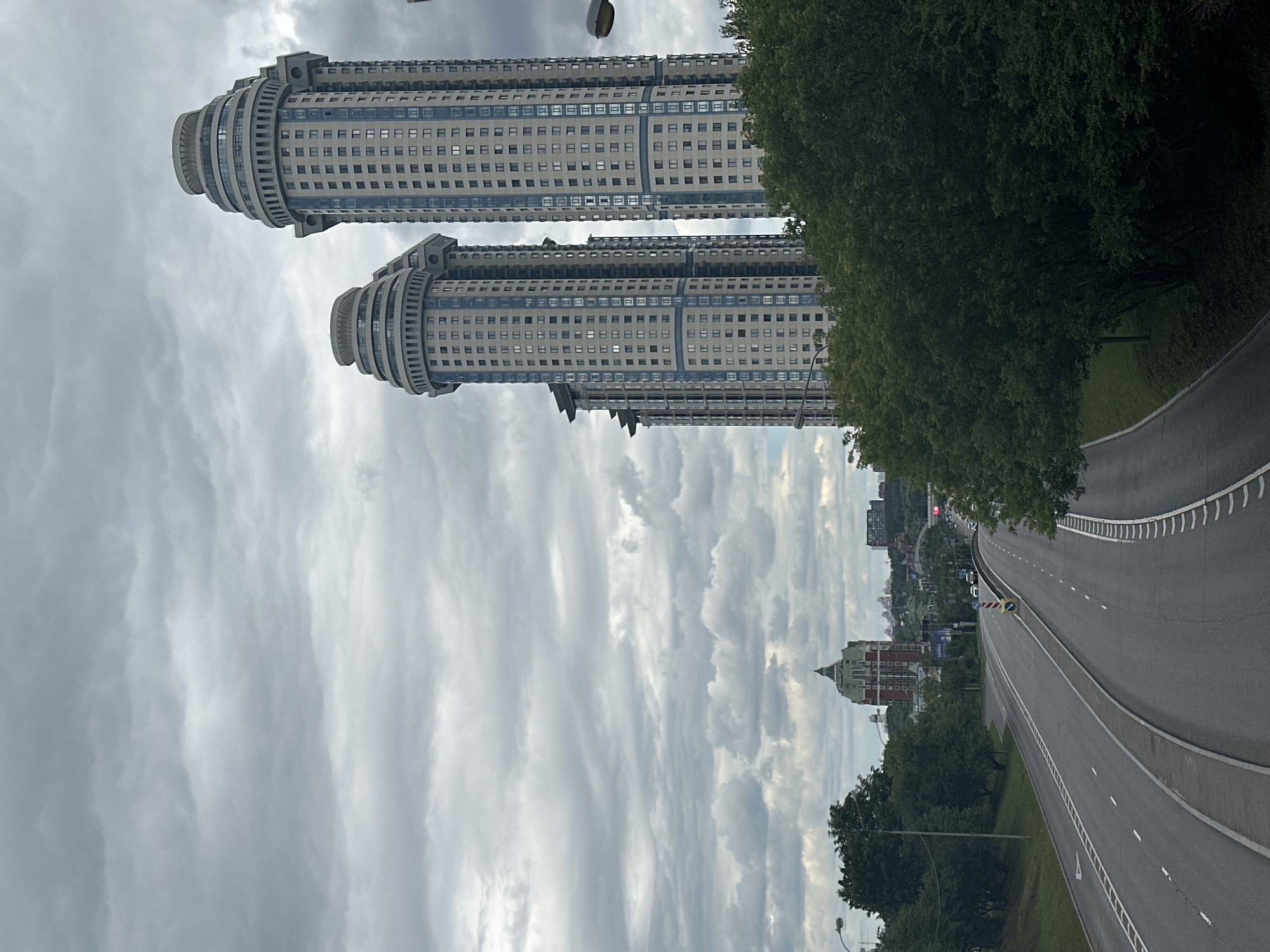 The image depicts a grey, cloudy sky over Moscow, Russia. Two identical, modern skyscrapers with rounded bases and light-colored facades accented with blue bands dominate the left side of the frame. The buildings are tall and appear to be residential or office complexes, featuring numerous windows. To the right, a multi-lane highway curves into the distance, bordered by a line of lush green trees. The highway has clear lane markings and white dashed lines. In the background, more buildings and trees are visible, suggesting an urban or suburban environment. The overall mood is overcast and somewhat subdued. The time of day appears to be daytime, but the cloudy weather diffuses the light. No people are clearly visible. There is a yellow and black traffic sign on the highway.