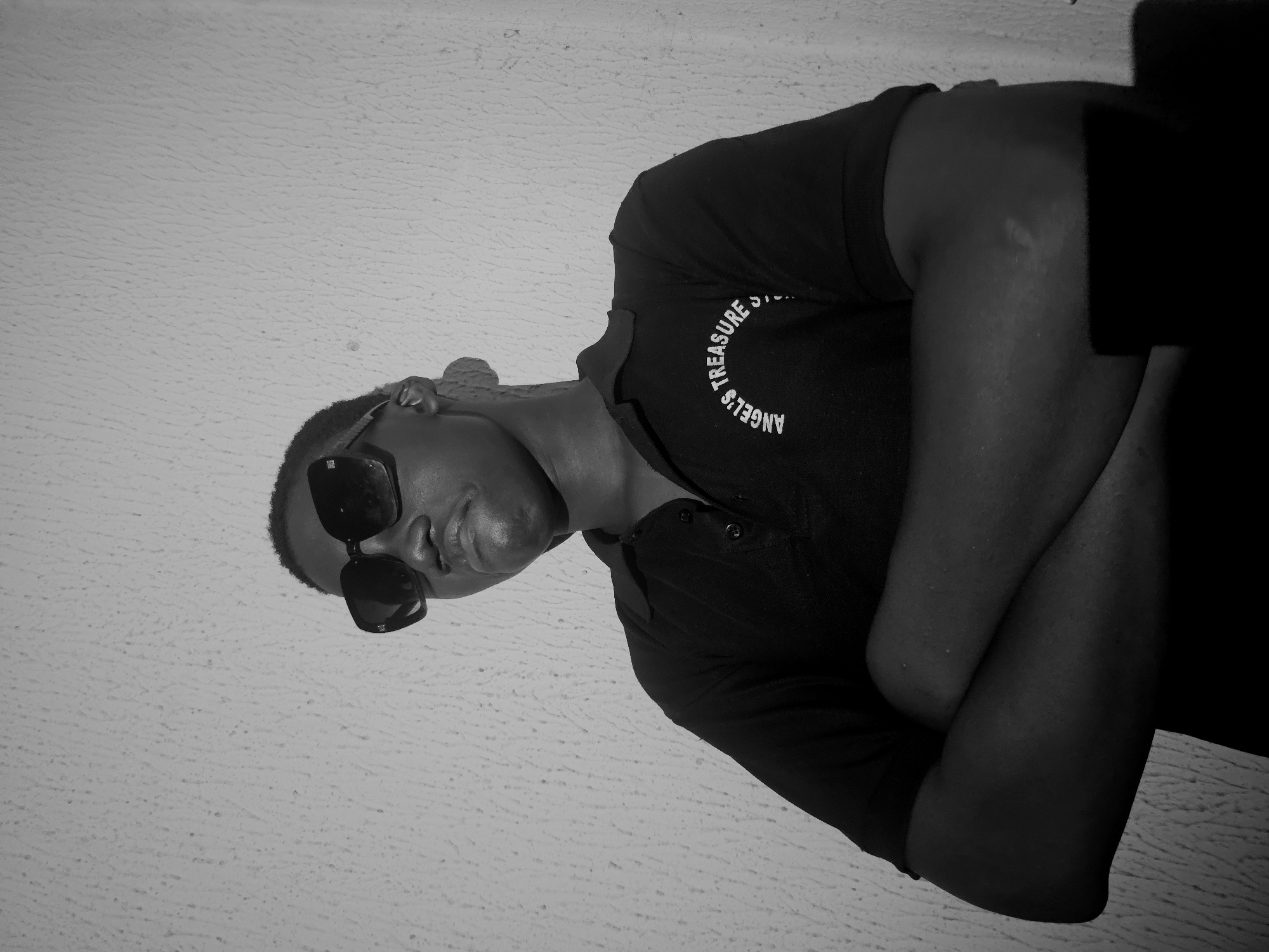 A monochrome image shows a man wearing sunglasses and a dark polo shirt with the words "ANGEL'S TREASURE S…" printed on it. He is standing with his arms crossed. The background appears to be a textured wall. The location and city are unspecified.