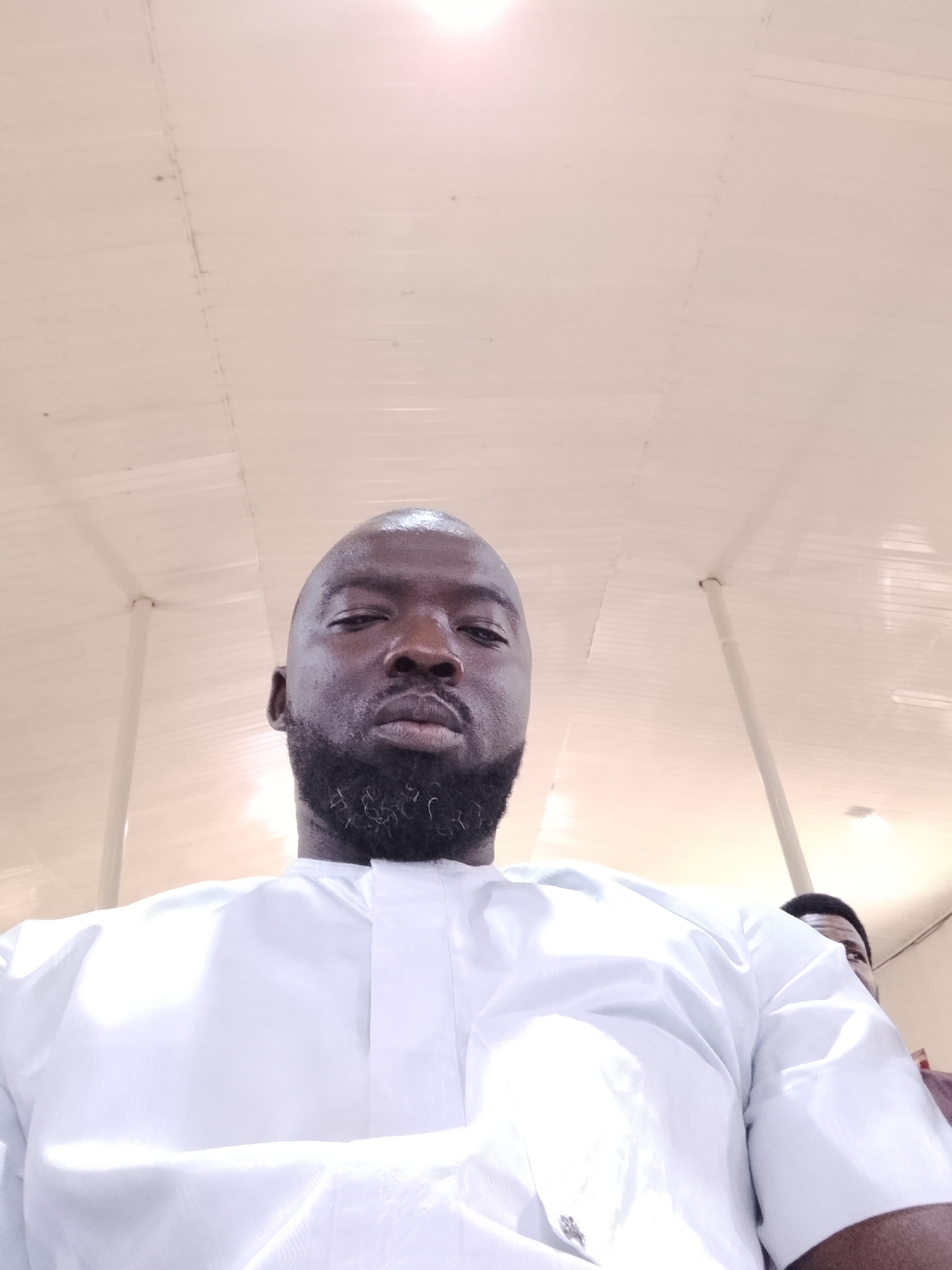A man with a dark complexion and a full beard is looking down at the camera. He is wearing a white shirt. Another person is partially visible in the background to the right of the frame. The background features a white ceiling with visible panels and structural supports. The location is in the city of Unknown, Unknown.