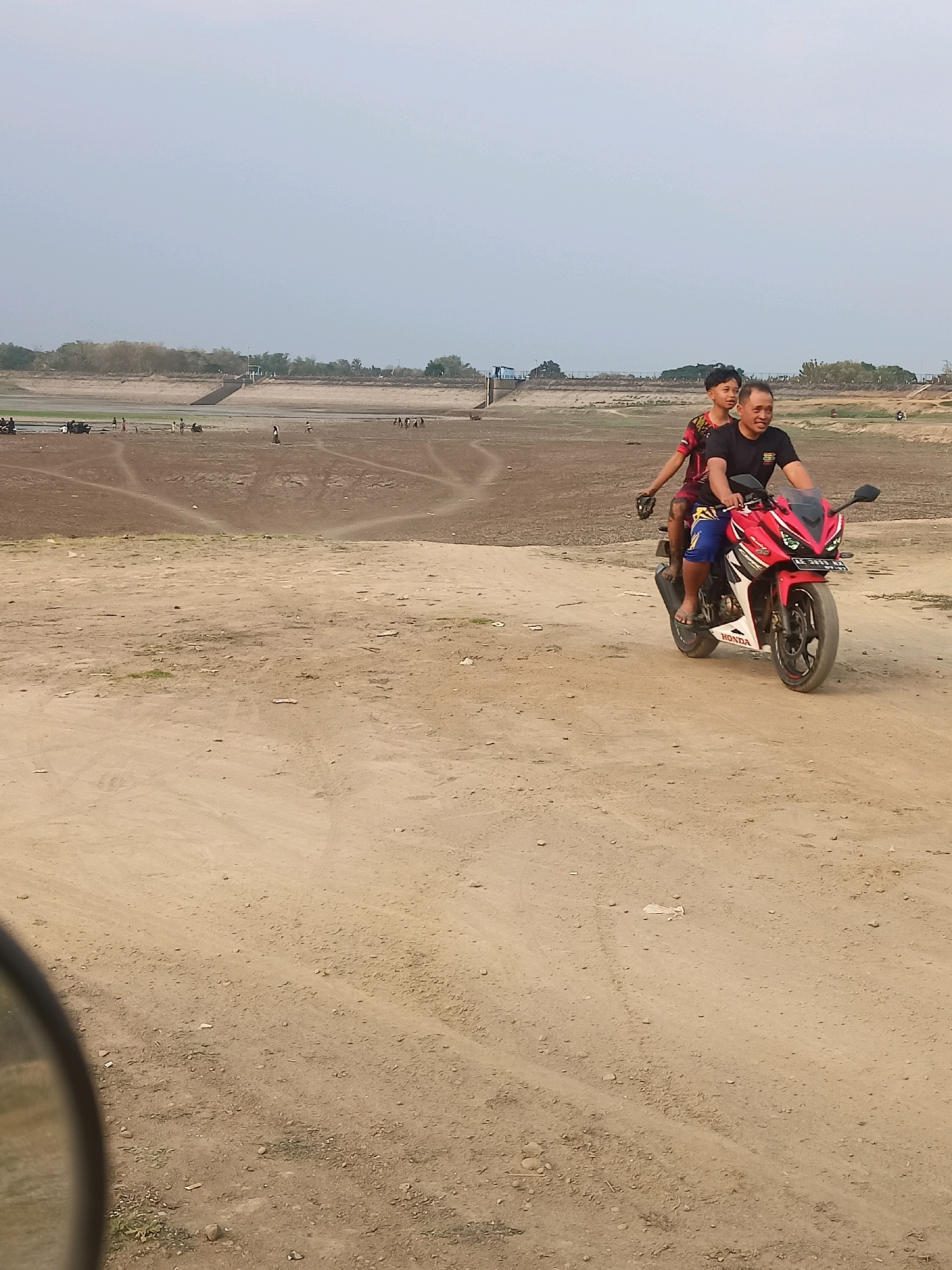 In Sambiduwur, Indonesia, a man and a young boy ride a red and white motorcycle on a dirt road. The man, wearing a black t-shirt and blue shorts, is driving, while the boy, in a red and black shirt, sits behind him. In the background, a dry, barren landscape stretches out, with a body of water visible in the distance, lined with trees and structures. Several other people and vehicles are scattered across the dry terrain. The sky is overcast, and the overall scene depicts a rural, possibly arid environment.