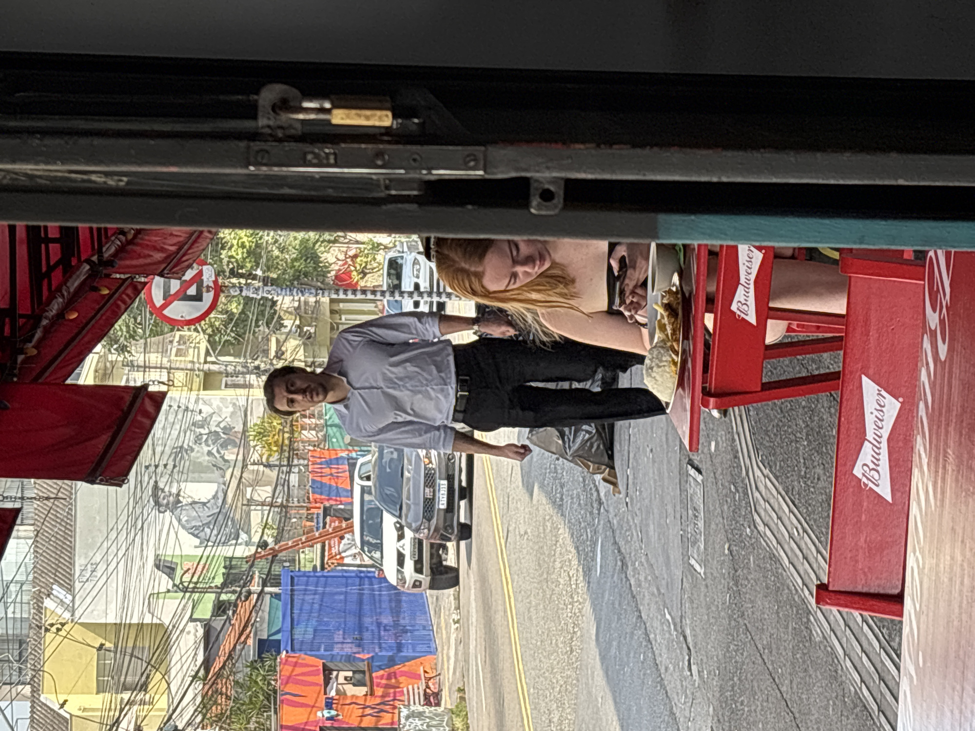 The image depicts a vibrant street scene in an urban environment, likely in São Paulo, Brazil, under bright daylight. In the immediate foreground, what appears to be an outdoor seating area of an establishment is visible. Two red chairs and a red table are prominent, with the "Budweiser" logo clearly emblazoned in white on the back of the chairs.
A woman with blonde hair is seated at the table, leaning forward and looking intently at a smartphone held in both hands. A plate of fried food, possibly fries or similar snacks, is visible on the table next to her. Standing nearby, facing the viewer with a slight smile, is a man dressed in a light-colored, collared shirt, black belt, and black trousers. He appears to be observing the surroundings.
Beyond the seating area, a paved street stretches into the distance, marked with a double yellow line. Several vehicles, including a white car, a silver SUV, and a blue truck, are parked or moving along the street. The background is characterized by a dense urban landscape, featuring buildings of varying heights and colors. A large, intricate mural or graffiti art covers a significant portion of one building's wall. Numerous overhead power lines and cables crisscross the scene, creating a characteristic urban visual texture. Red awnings are also visible on buildings to the left.
A circular regulatory sign, red with a white background and a diagonal line, similar to a "no entry" sign, is partially visible. The bright sunlight casts distinct shadows, indicating a clear, sunny day. The overall impression is a bustling city street with a casual outdoor dining or social setting. A padlock is visible on a dark metal structure at the top of the frame, suggesting the photo was taken from within a window or doorway.