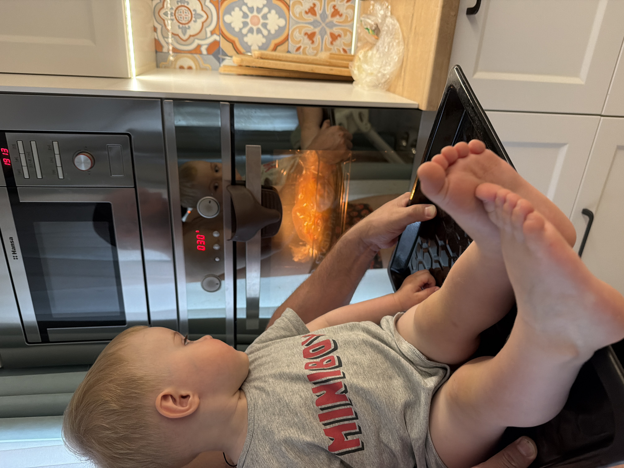 The image depicts a young child lying on their back in a black baking tray held by an adult. The child is wearing a gray T-shirt with the word "MINIBOSS" in red and black lettering. The focus is on the child's feet, which are close to the camera.
In the background, an oven is visible, with its door reflecting the image of the person holding the baking tray and what appears to be food inside. The oven has a digital display showing "19:13" and "030." Above the oven is a countertop with various items, including a stack of wooden boards and a bag. White cabinets with dark handles are also visible in the background.
There is no indication of Votkinsk, Russia.