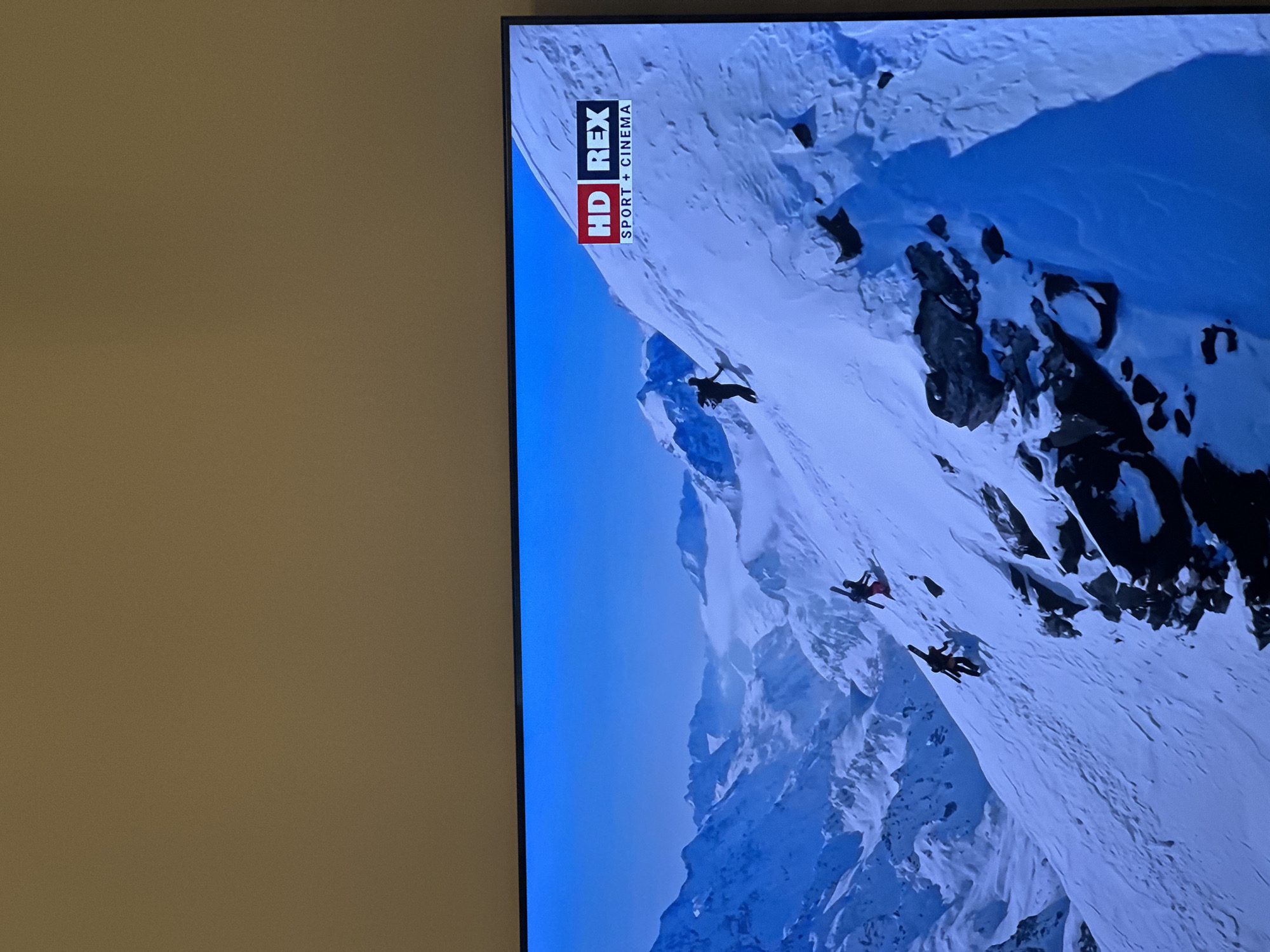 An indoor photograph taken in Kharnavy, Russia, shows a television screen displaying a mountainous winter landscape. The upper portion of the image captures a plain, light brown wall. The television screen, framed by a dark bezel, occupies the lower part of the frame. On the screen, a vast expanse of snow-covered mountains is visible under a clear blue sky, with a layer of clouds or fog settled in the distant valleys. Three human figures, appearing as dark silhouettes, are actively ascending a steep, snow-covered slope in the foreground. Each figure carries skis or similar equipment strapped to their backs. Dark rock outcrops are interspersed within the snow on the slope. A red and white channel logo, "HD REX SPORT + CINEMA," is visible in the upper right corner of the television display.