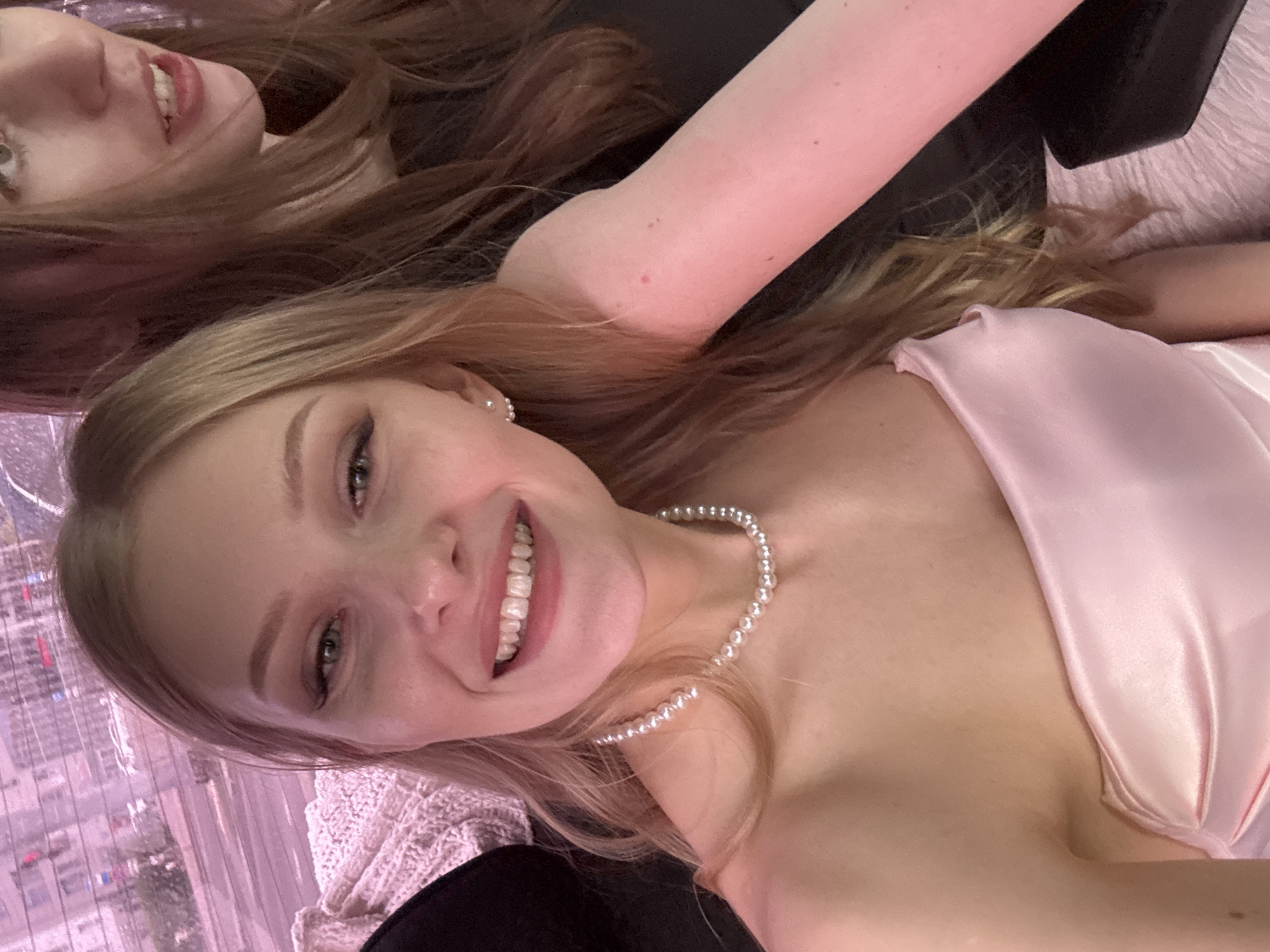 Two women are in a photograph, likely a selfie. The woman in the foreground, with blonde hair, is smiling and wearing a pearl necklace and a light pink top. She is facing the camera. To the upper left, another woman with reddish-brown hair is visible, partially cut off in the frame. Outside a window behind them, a cityscape is visible, featuring buildings, potentially in Perm, Russia.
