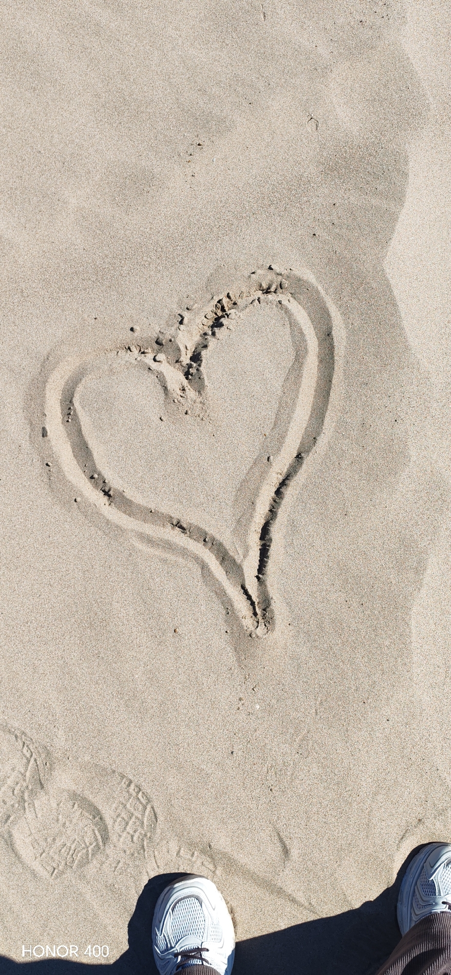 A downward-angle, ground-level shot depicts a heart drawn in sand. The sand is light brown and textured. The heart is centered, and the lines forming it are darker than the surrounding sand due to being dug into it. A pair of sneakers, primarily white with visible laces, are positioned at the bottom of the frame. The bottom left sneaker partially obscures the bottom left corner, and the bottom right sneaker does the same for the bottom right corner. A footprint is faintly visible in the sand to the left of the left sneaker. The lower left corner has "HONOR 400" displayed in small, black text. The location is Noviy, Russia, implied as being a beach or sandy area. No people are fully visible in the frame, only partially visible footwear. No interaction is observed.
