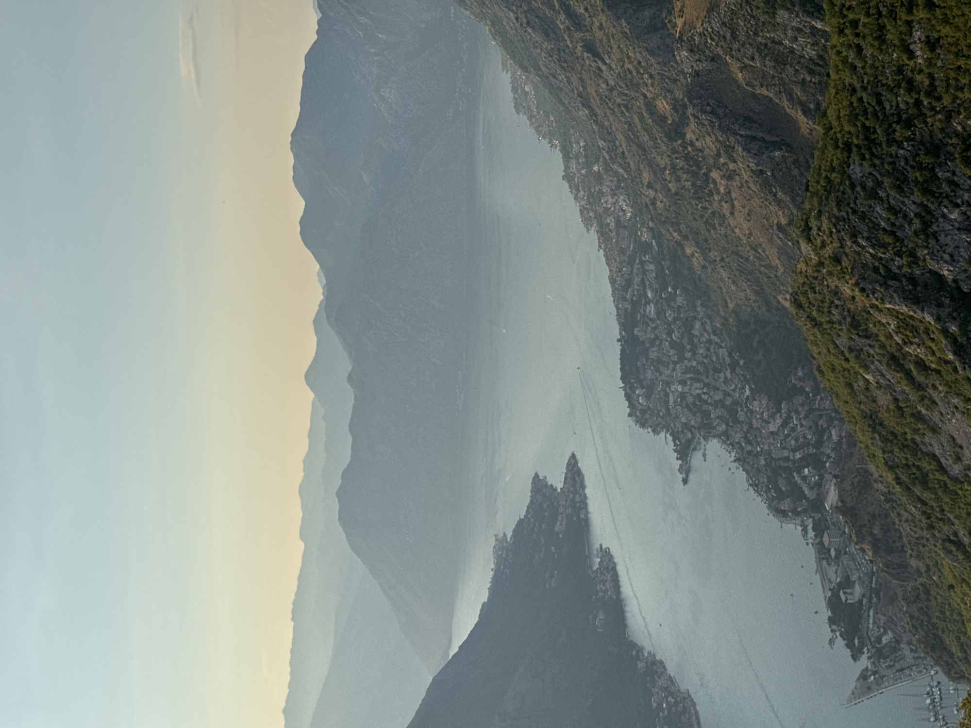 The image is an aerial view of the Bay of Kotor, specifically showing the coastline near the town of Žanjev Do, Montenegro. The scene captures a wide expanse of water, bordered by steep, mountainous terrain. On the western side of the bay, a densely populated settlement with numerous buildings stretches along the shoreline. The water is a light blue-gray, with several small boats visible, leaving subtle wakes. The mountains are covered in scrub and sparse vegetation, with visible rock faces. In the background, successive layers of mountains fade into a hazy, pale blue sky, suggesting a clear, possibly warm day. The light suggests it might be late afternoon or early morning, with a gentle gradient of color in the sky from pale yellow near the horizon to blue above. The perspective is from a high vantage point, looking down and across the bay.