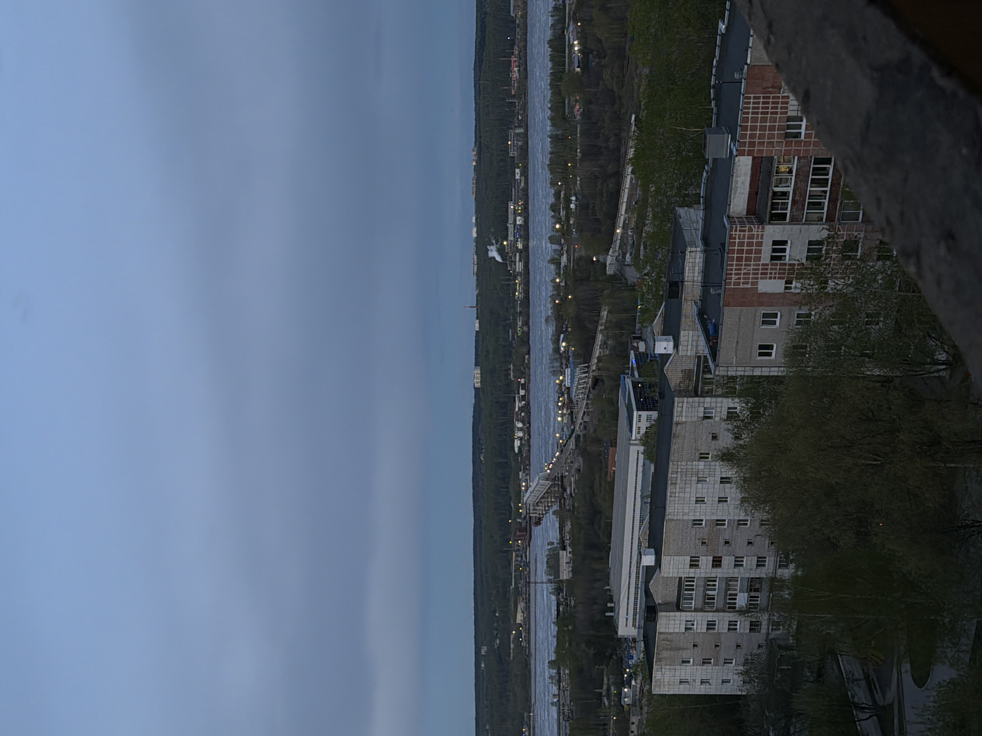 The image depicts a nighttime urban scene in Perm, Russia, viewed from a high vantage point, possibly a balcony. A wide river bisects the frame vertically, with illuminated buildings and streetlights visible along its banks. Dense forest lines one side of the river. Residential buildings with visible windows are clustered on the right, partially obscured by trees and architectural elements. The sky is overcast and dusky. A bridge spans the river in the midground.