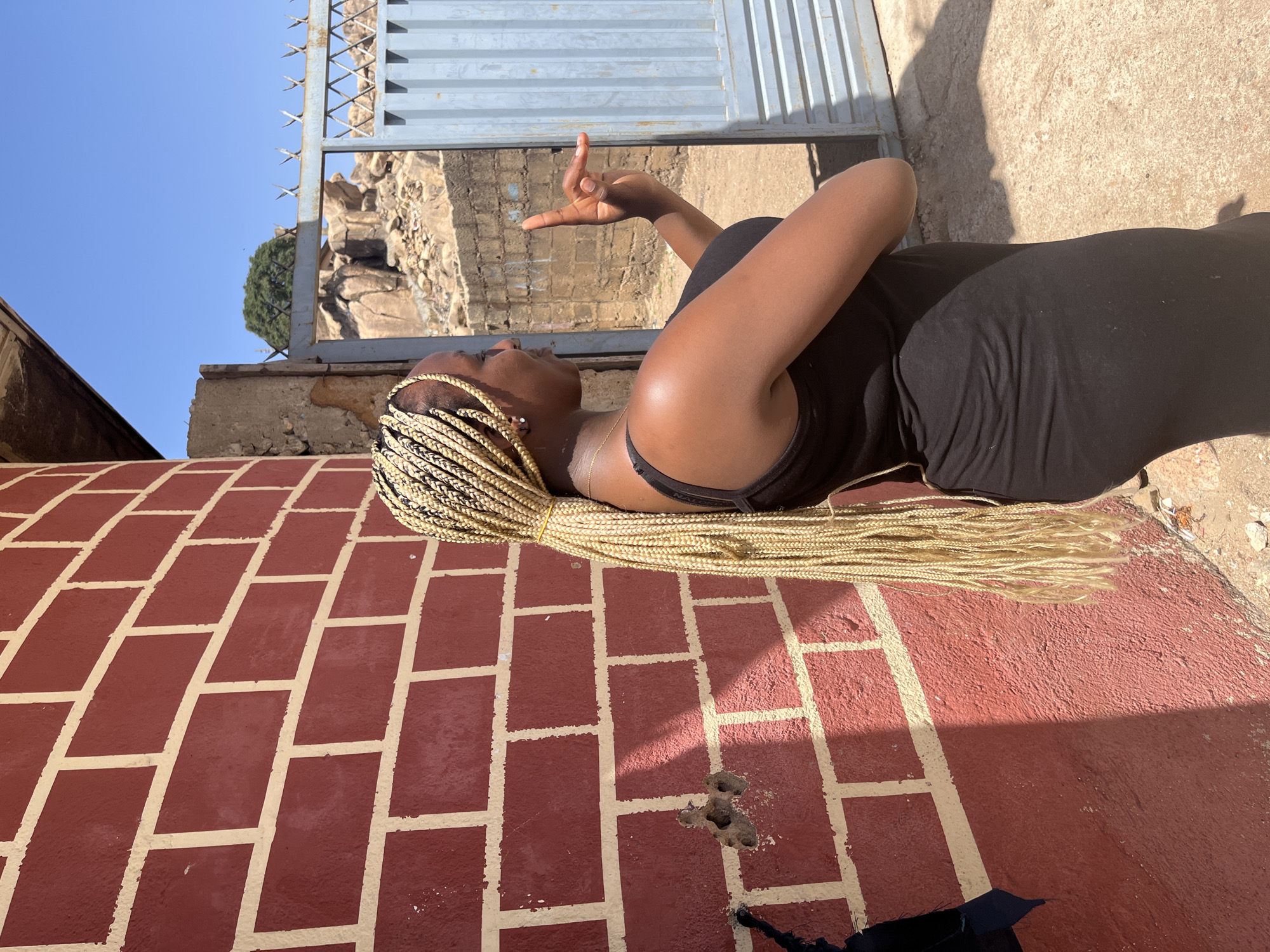A woman with blonde braided hair is lying on her side on a red brick surface with light grout lines in an outdoor setting. She is wearing a black sleeveless dress. Her head is turned upwards and her right arm is raised with her fingers extended. In the background, there is a light blue gate, beyond which a rocky cliff or quarry is visible. The sky is clear and blue. The location appears to be Unknown, Unknown.