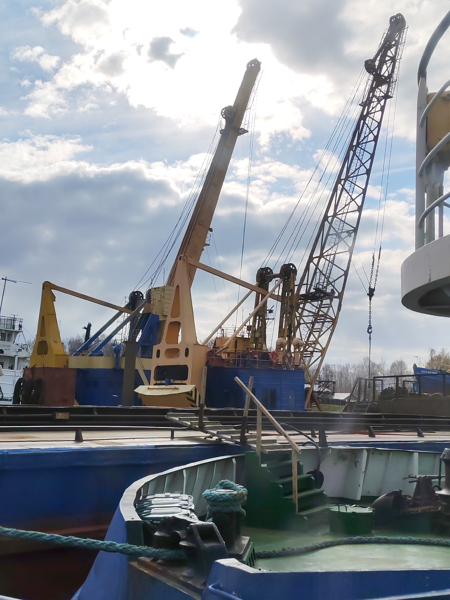 The image captures a maritime industrial scene, likely a port or shipyard, featuring two large cranes positioned on a blue and yellow barge or specialized vessel, with other boats docked nearby. In the foreground, the bow of a blue boat with a green deck is visible, featuring a thick green mooring rope secured to a bollard, a set of wooden stairs, and various deck fittings.
In the mid-ground, the two cranes dominate the scene. The crane on the right has a prominent lattice boom structure, with its hook and chain dangling. The crane on the left has a solid-looking yellow boom. Both cranes exhibit complex pulley systems and cables. Their main bodies are a mix of yellow and blue, suggesting they are part of a floating crane operation.
Behind the cranes, another white boat is partially visible on the far left, bearing Cyrillic text on its hull: "БД-0401 ЗОН-503" and "БД-0401 ЗОН". In the distance, a strip of land with trees can be seen.
The sky is partly cloudy with bright sunlight, which is partially obscured by clouds and creates a backlit effect for the cranes, especially towards the upper right, indicating it is daytime. The overall impression is one of heavy industrial equipment in a working port environment.