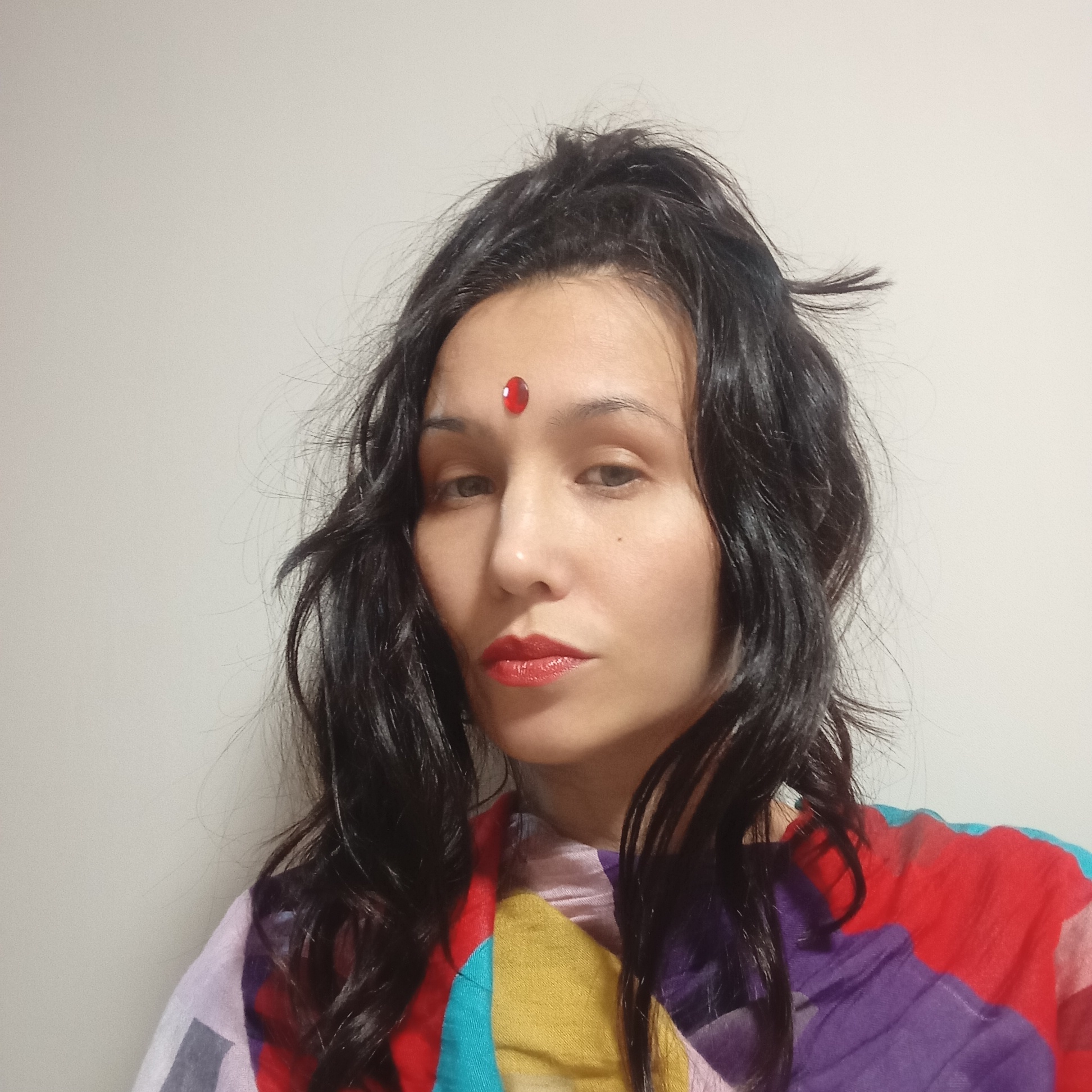 This is a medium close-up portrait of a woman, framed against a plain, light-colored wall, suggesting an indoor setting in Pervomaysky, Russia. The woman has long, dark, wavy hair, appearing somewhat dishevelled, which falls around her face and shoulders. She wears a vibrant red bindi on her forehead and red lipstick, indicating cultural adornment. Her eyes are dark, and her expression is neutral to slightly contemplative, looking directly at the viewer. She is dressed in a colorful garment, possibly a scarf or shawl, featuring a patchwork or abstract design with bold blocks of red, purple, yellow, and blue, interspersed with lighter tones. The lighting is soft and even, without harsh shadows. No specific activity is taking place beyond posing for the photograph. There is no visible text in the image.