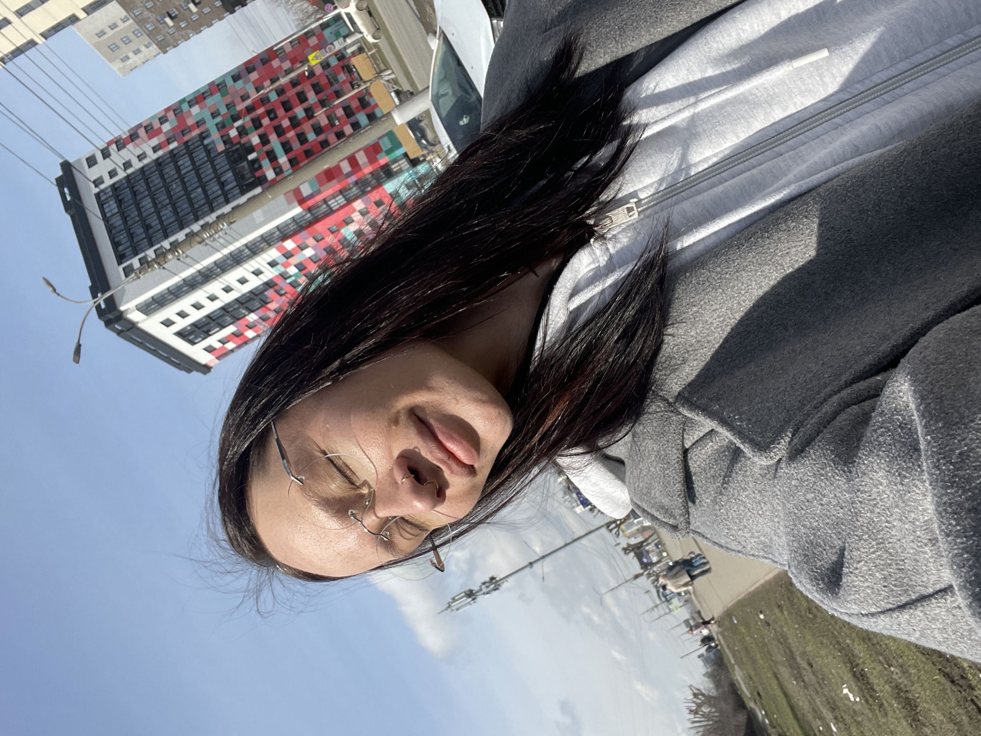 A woman wearing glasses, a white zippered hooded sweatshirt, and a gray coat takes a selfie in front of a tall building with a red, white, green, and black geometric pattern in Izhevsk, Russia. The sky is blue with some clouds. A street with cars and utility poles is visible in the background.