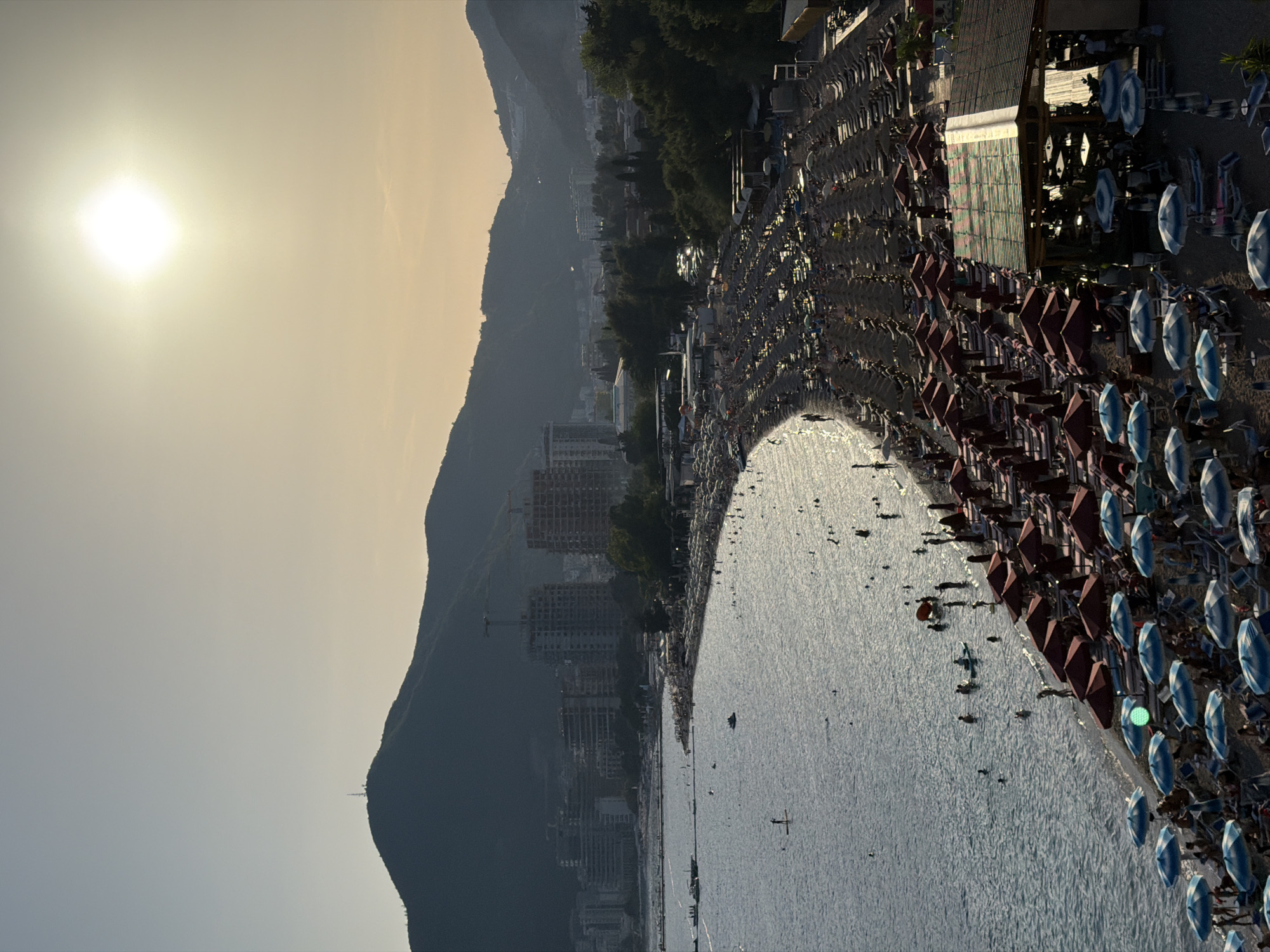 The image captures a bustling beach scene in Boreti, Montenegro, during what appears to be late afternoon or early evening. The sun is a bright, hazy disc in the upper right portion of the sky, casting a warm, golden light. In the foreground, a long stretch of sandy beach is densely populated with rows of lounge chairs and umbrellas. Many of these umbrellas are a deep red, while others are blue and white striped, providing shade for beachgoers.
The water, a shimmering blue, laps gently at the shoreline. Scattered throughout the water are numerous people swimming, wading, and engaging in water activities. Several individuals are seen on paddleboards, one standing upright. Small boats are anchored further out in the bay.
Along the edge of the beach, there are structures including a beach bar with a distinctive patterned roof, and various beach amenities. Further inland, the landscape is dominated by the silhouettes of mountains, with the outlines of buildings and construction cranes visible on the slopes, indicating urban development. The overall impression is one of a vibrant, popular tourist destination enjoying the last hours of daylight.