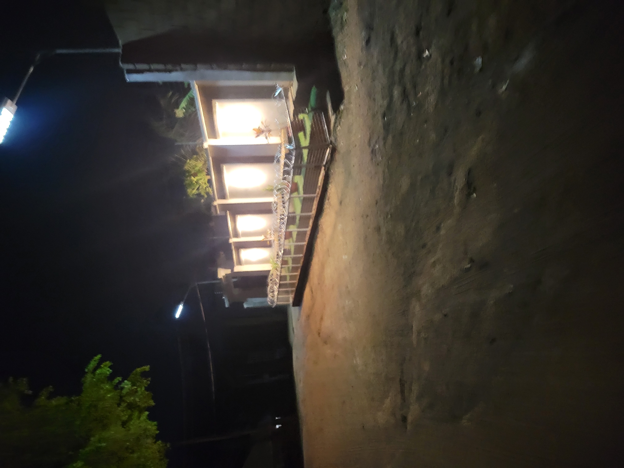 A nighttime image shows a multi-story building with evenly spaced, illuminated windows. A lamppost illuminates the building and nearby tree. The environment consists of a dark surface and what appears to be ground cover, possibly soil. The image is rotated. The location is suggested to be Jos, Nigeria.