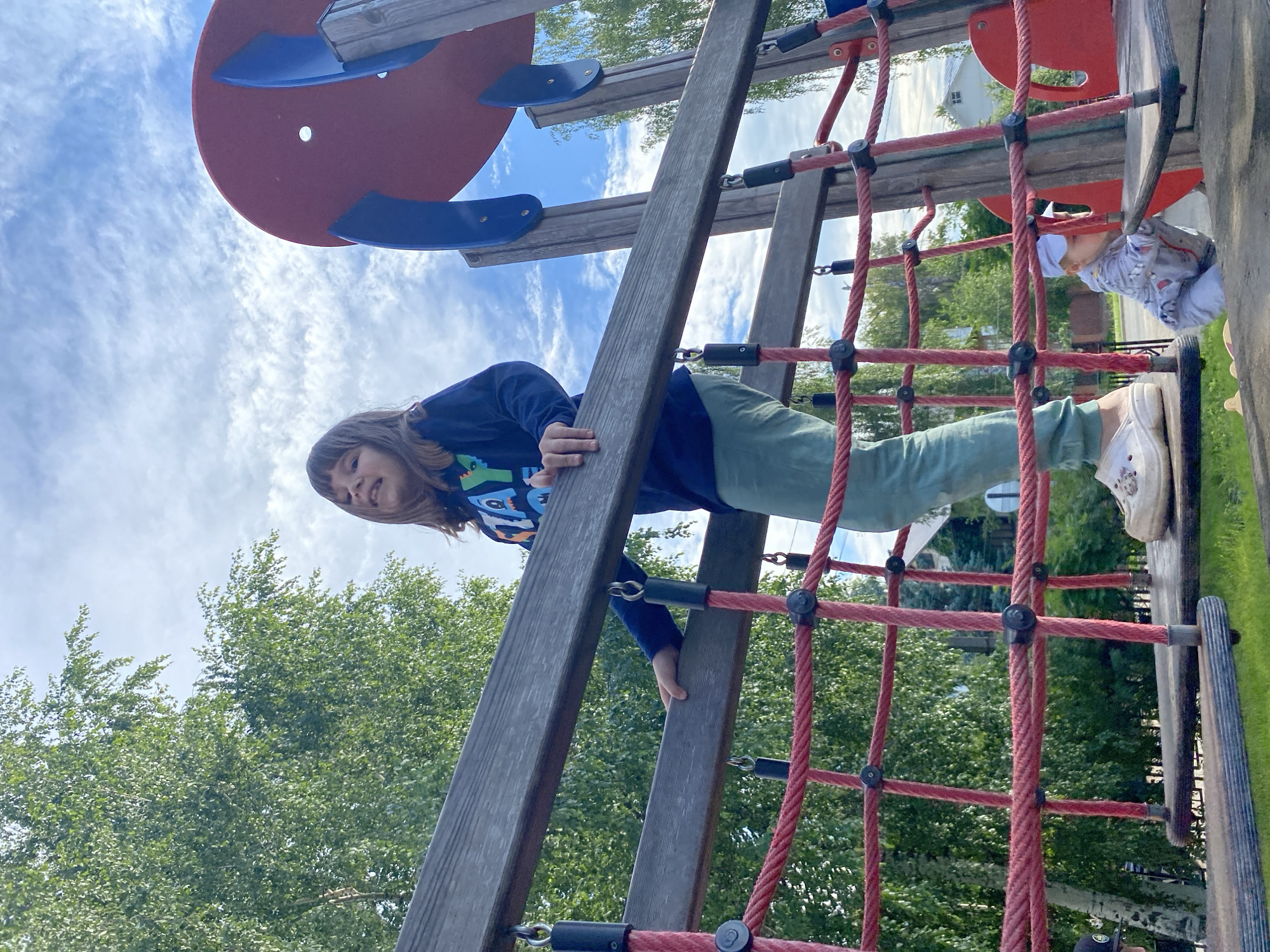 A low-angle outdoor shot captures a young girl standing on a wooden platform of a playground, her body angled towards the camera. She is wearing a dark long-sleeved shirt with a graphic design and light-colored pants, her right hand resting on a wooden beam above her. Her face is turned towards the viewer, and she appears to be smiling or making a face. Below her, on a lower platform, a baby in a light-colored outfit and hat is visible, reaching for something. The playground structure is made of wood and red rope netting. In the background, green trees and a blue sky with white clouds are present. The scene is set in a park or recreational area within the city of Chaikovsky, Russia.