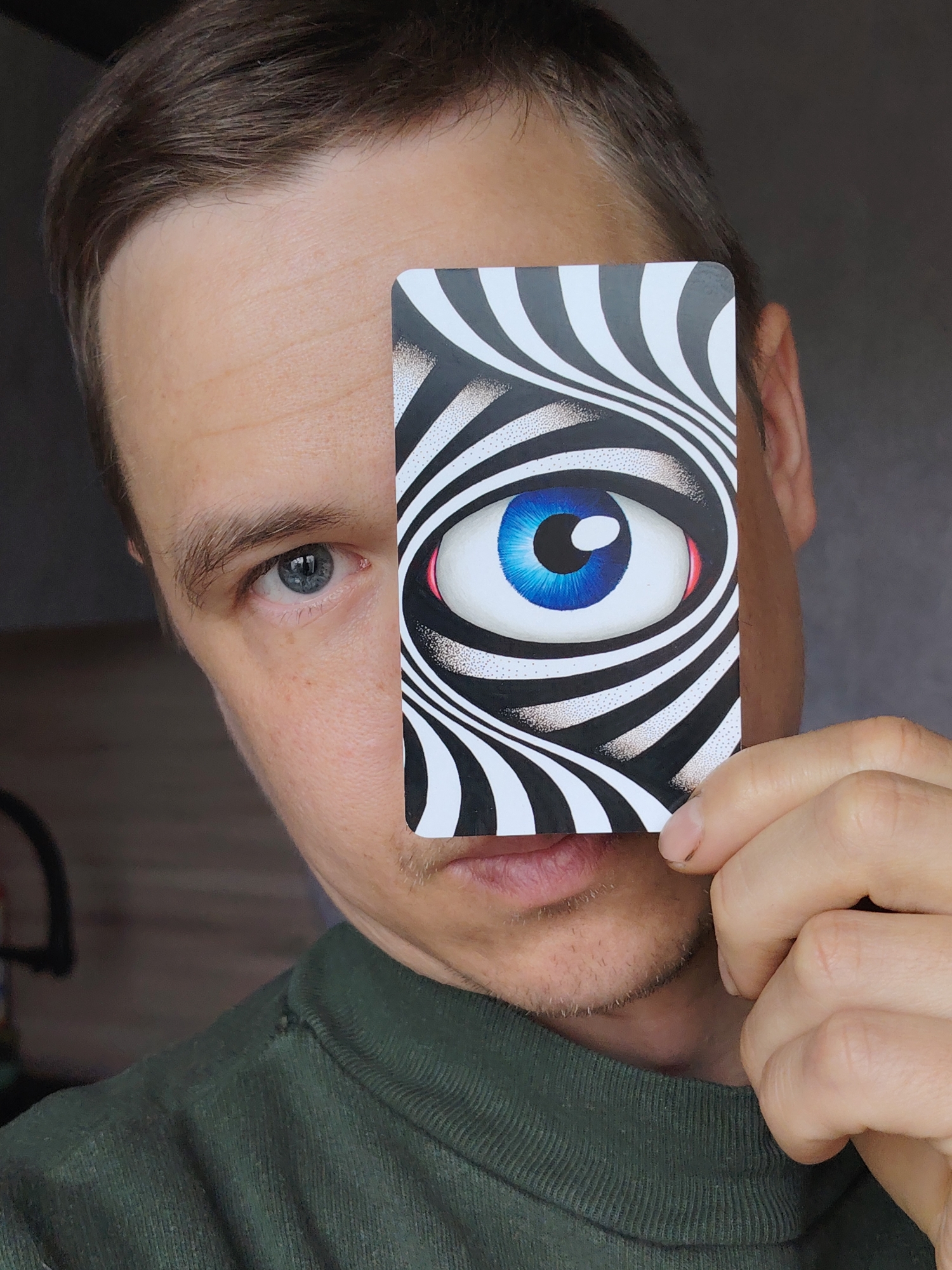 A mid-shot portrait features a man holding a rectangular card over his right eye, taken indoors in Chaikovsky, Russia. The man has short brown hair, fair skin, and light stubble on his chin and upper lip. His visible left eye (viewer's right) is blue-gray and appears to hold a neutral expression. He is wearing a dark green, ribbed long-sleeved shirt or sweater. His left hand (viewer's right) holds the card vertically, covering the right side of his face from just above the eyebrow to below the mustache.
The card's design depicts a stylized eye with a vibrant blue iris and black pupil, surrounded by a subtle red detail near the tear duct area. This eye is set against a background of swirling black and white stripes, creating an optical illusion effect. The background behind the man is softly blurred, showing muted dark tones consistent with an indoor setting, possibly a wall or interior room. Part of a dark, out-of-focus object or furniture is visible on the left side of the frame. The lighting is even, suggesting either natural indirect light or artificial illumination. The man is clearly posing for the photograph, using the card as a central visual element. There is no visible text in the image.