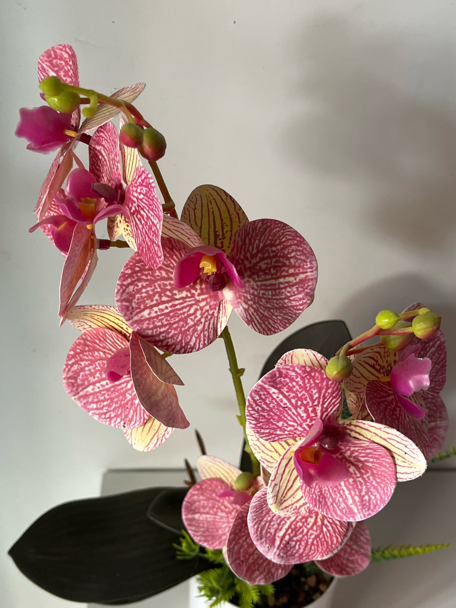 A close-up, angled shot features a spray of artificial pink and white orchids with yellow accents against a plain, light-colored wall. The orchids are arranged on a stem, with some flowers in full bloom and others as buds. Some dark, artificial leaves are visible at the bottom of the frame, partially obscuring a white pot. The lighting suggests an indoor setting. There is no information in the image to indicate the location of Tungkaran Pangeran, Indonesia.