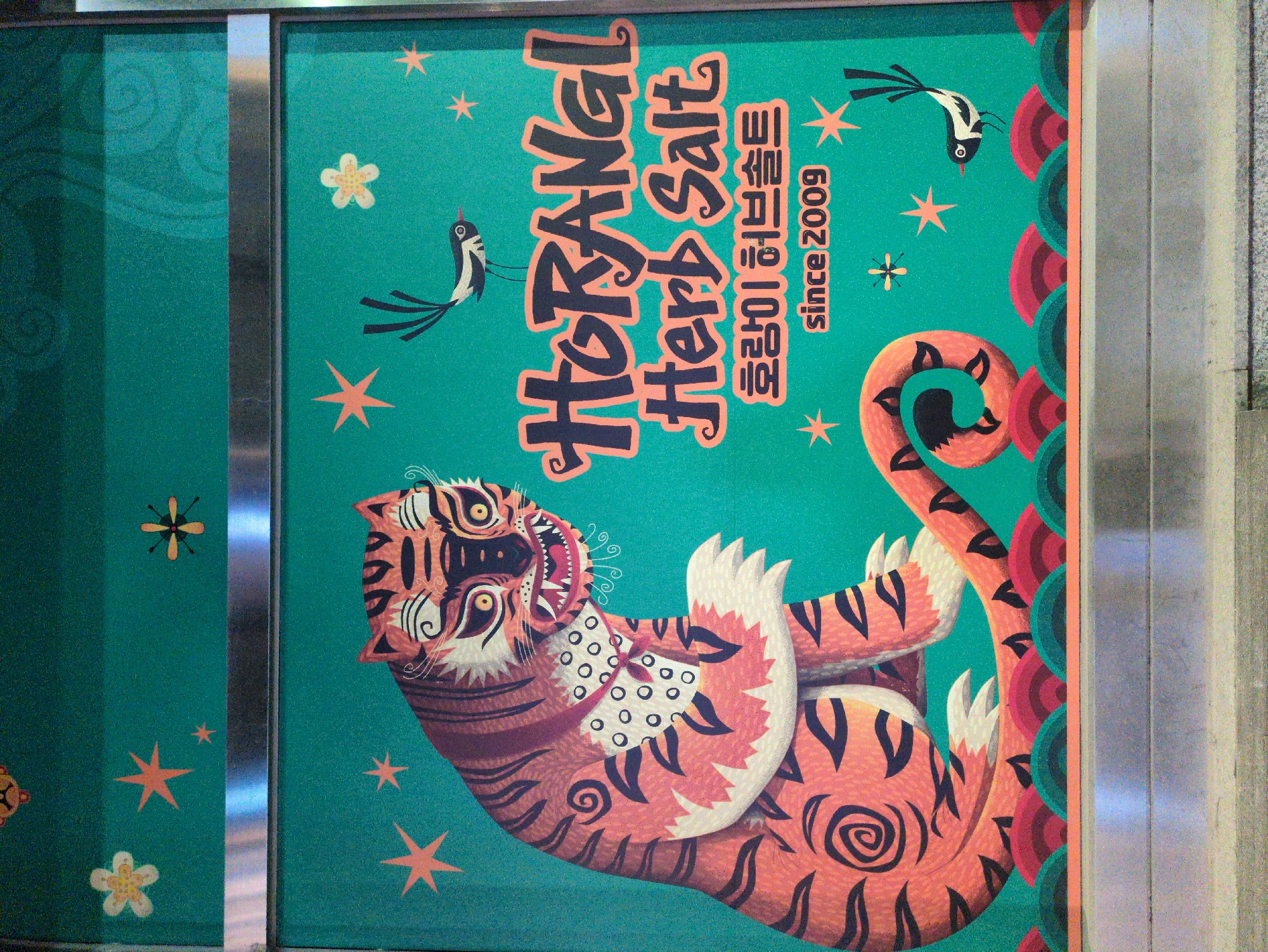 The image depicts a sign or mural, likely found in Seoul, South Korea, featuring a stylized tiger and birds on a green background. The tiger is drawn in a traditional folk art style, with orange and brown stripes, a white mane, and a fierce expression. Two black and white birds, possibly magpies, are positioned above the tiger. Text in Korean and English reads "HorangI Herb Salt since 2009". The mural is framed by metal or steel edges. The setting appears to be indoors, possibly a subway station or public space.